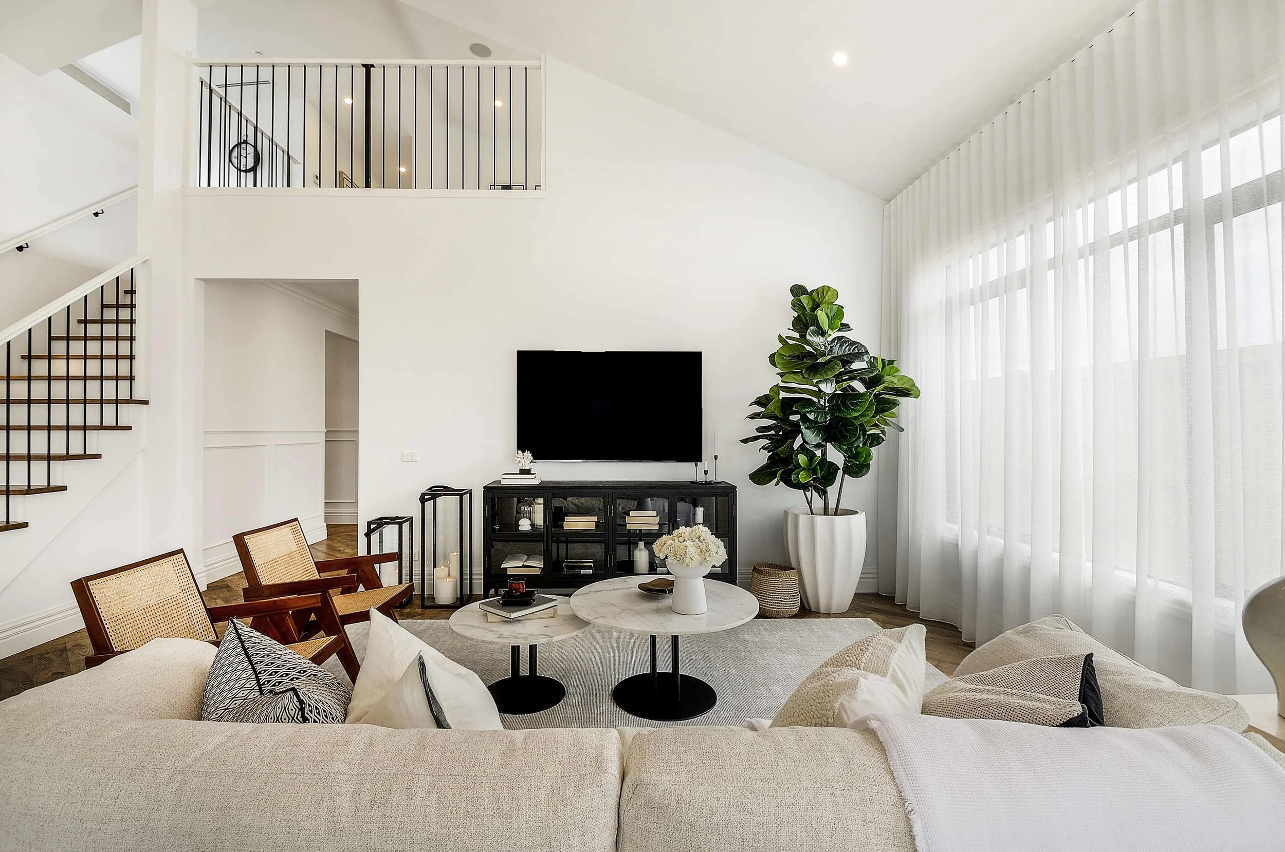 Bright living room with white walls and large window with sheer curtains.  There is a staircase to the left leading to an attic retreat that overlooks the open living area.
