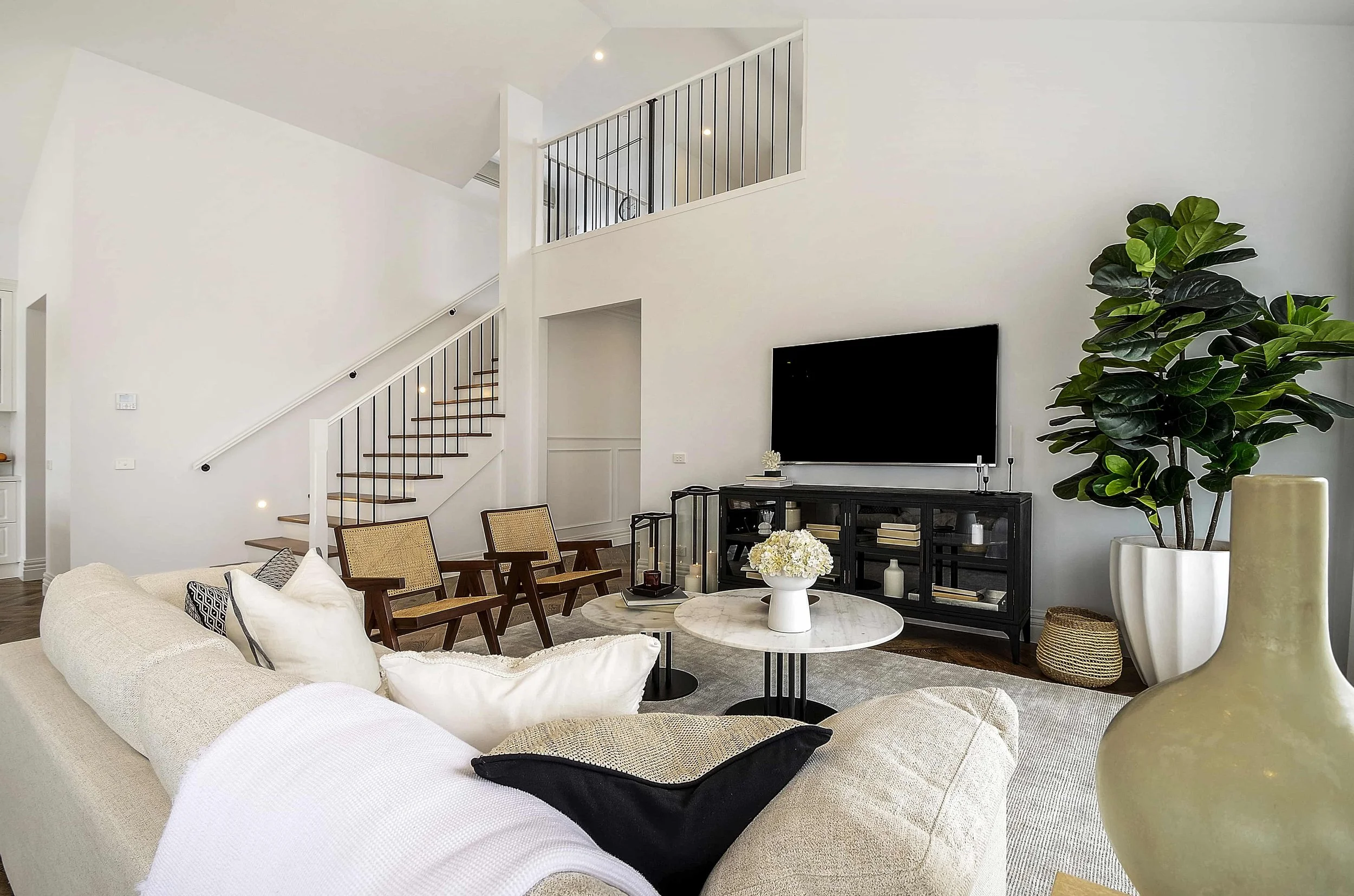 Modern living room with white walls, staircase with black railings, and a large flat-screen TV on a black console. There are various furniture pieces including a white sofa, a dining table with chairs, and decorative items like plants and flowers.