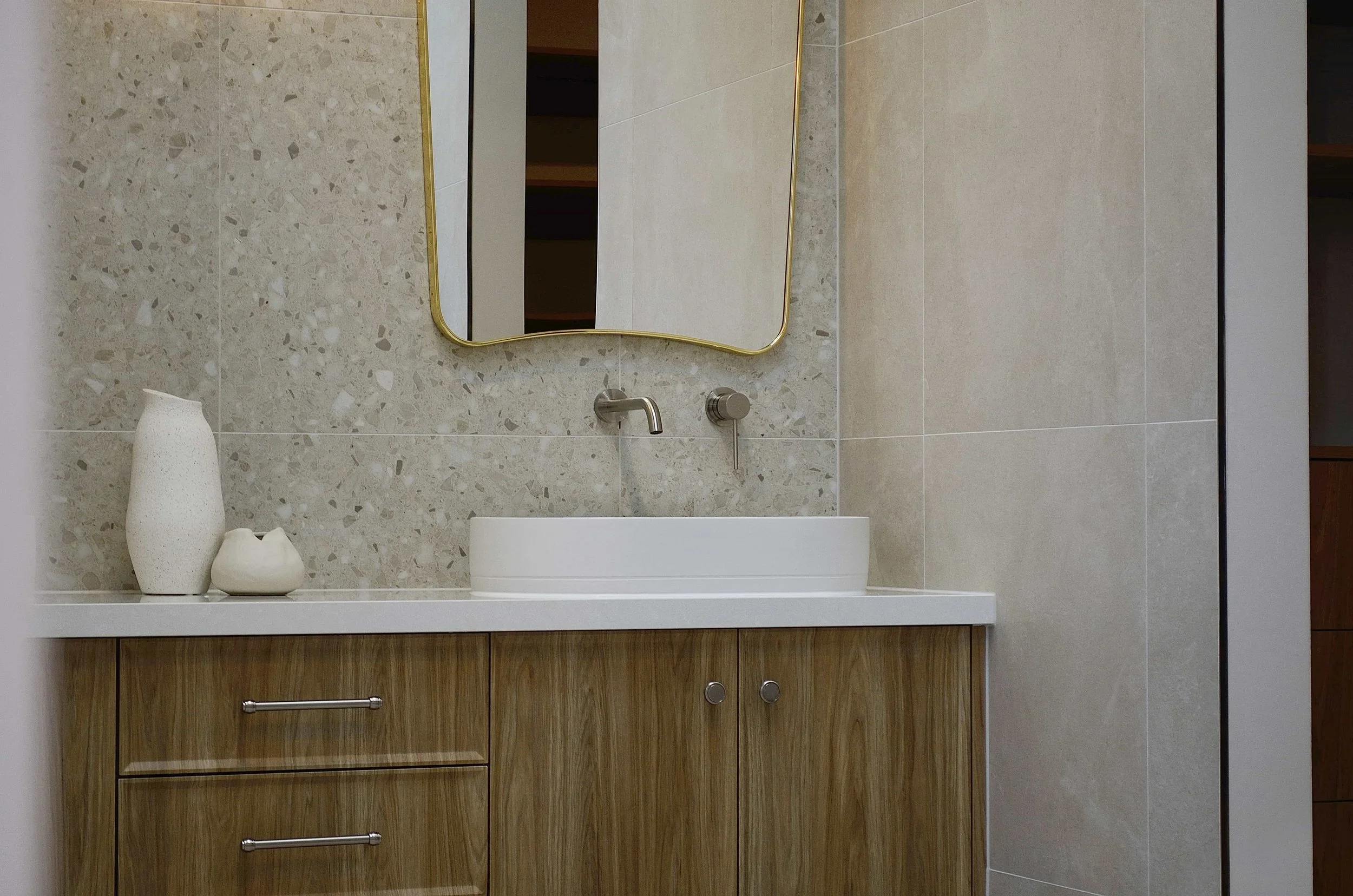 Modern bathroom vanity with timber doors, a white above bench oval basin, a golden-framed mirror, and minimal decorative vases in white, against beige and terrazzo tiled walls.
