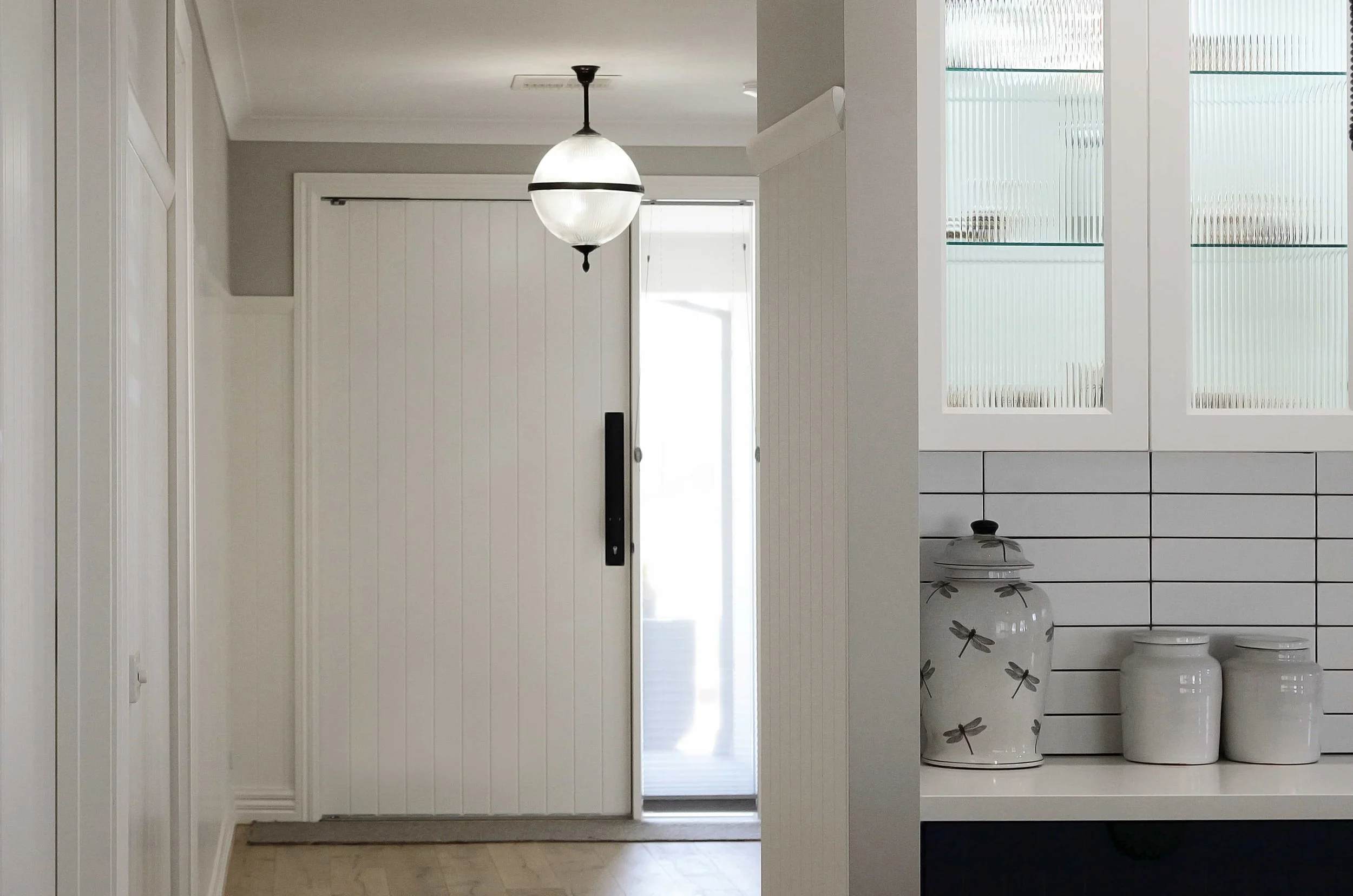 Interior view of a modern home entryway with a hanging pendant light, a large entry door, and overhead kitchen cabinets with fluted glass doors.