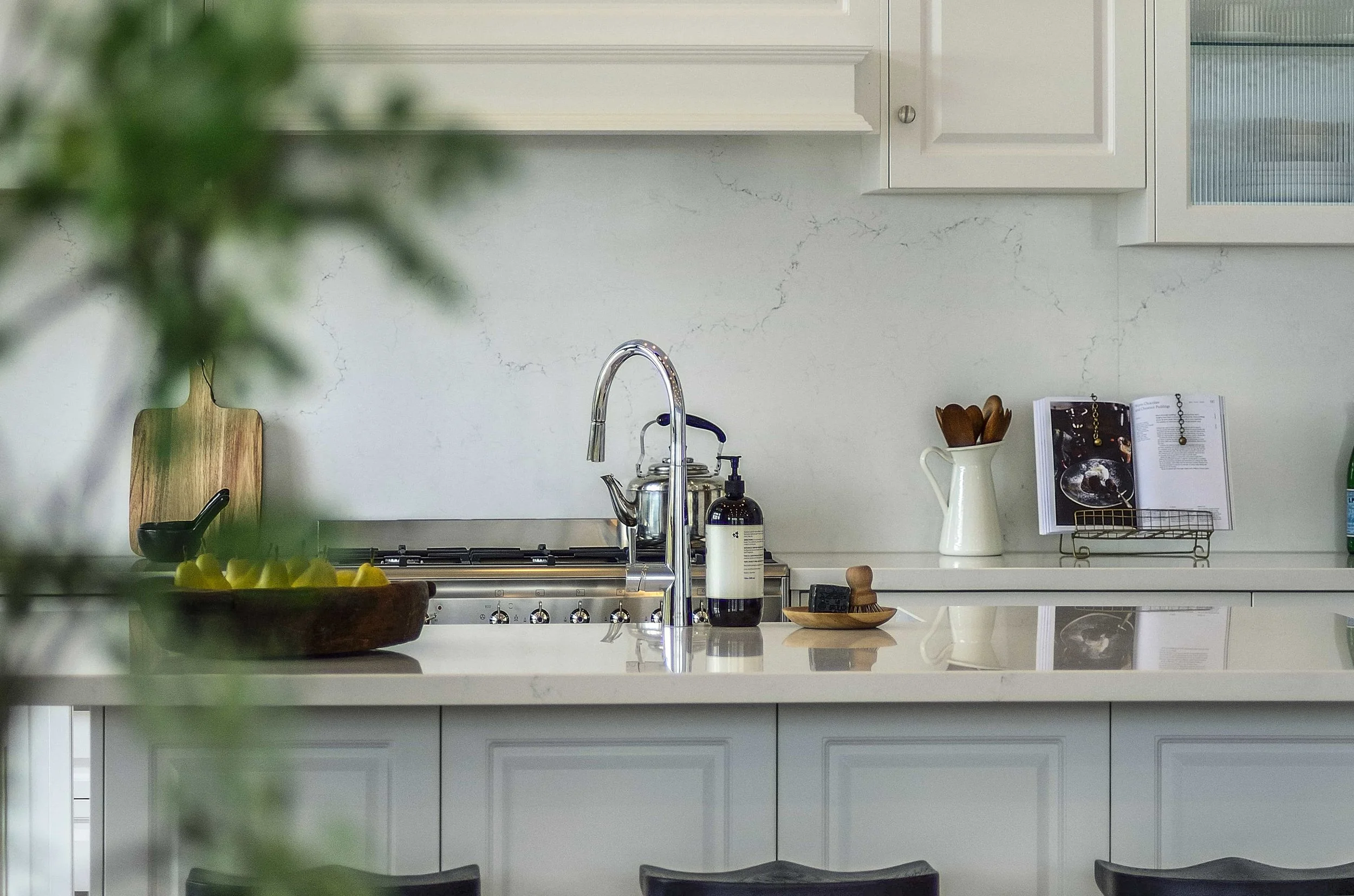 A modern kitchen with a white marble benchtop, routed door panelling, and a chrome faucet. The feature splashback is also in a natural marble stone. Decorative items are on the kitchen bench.