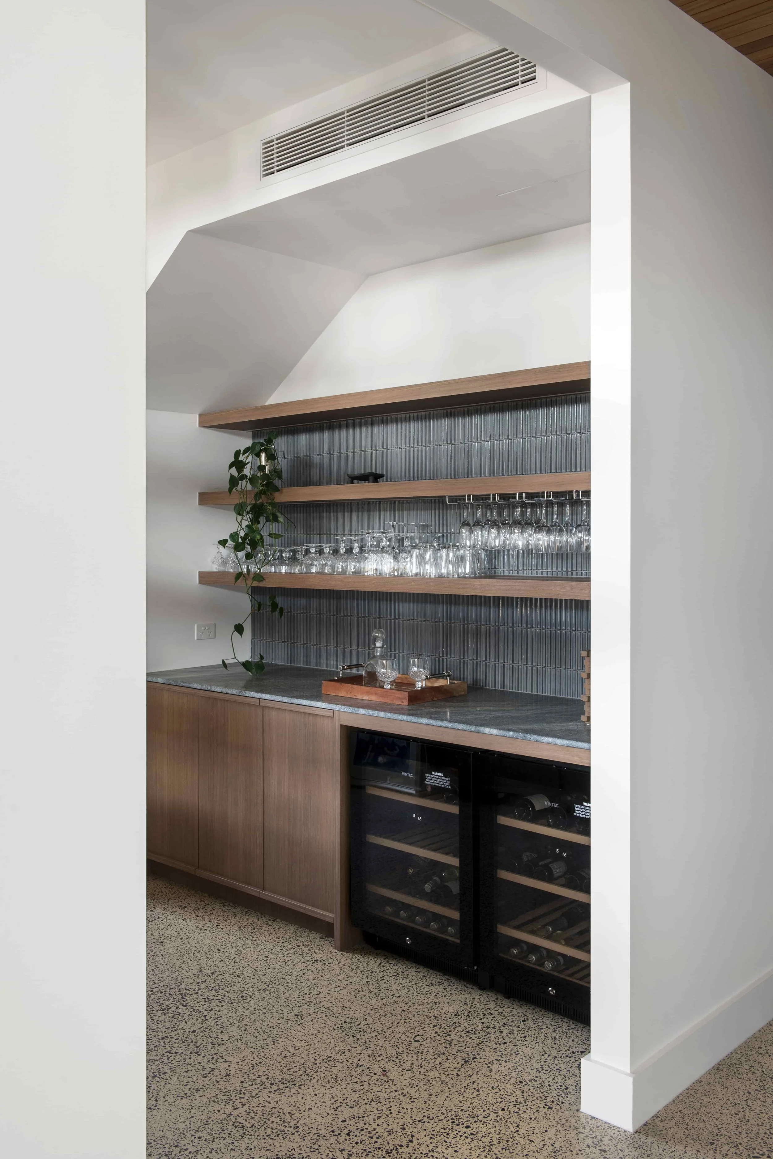 Modern kitchen corner with floating wooden shelves, a plant, and a wine cooler, featuring a speckled tile floor and minimalist design.