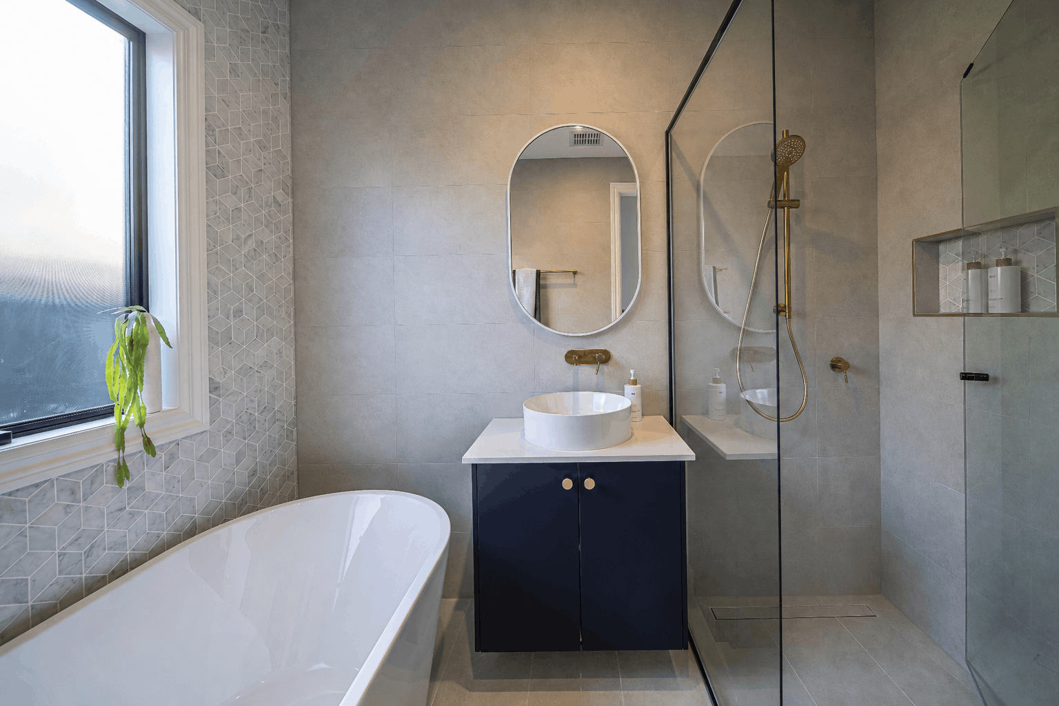 Modern bathroom with a bathtub near a window, a round basin with a mirror above, and a walk-in shower with gold plated fixtures and a glass door. There is a feature hexagonal tile behind the bathtub.
