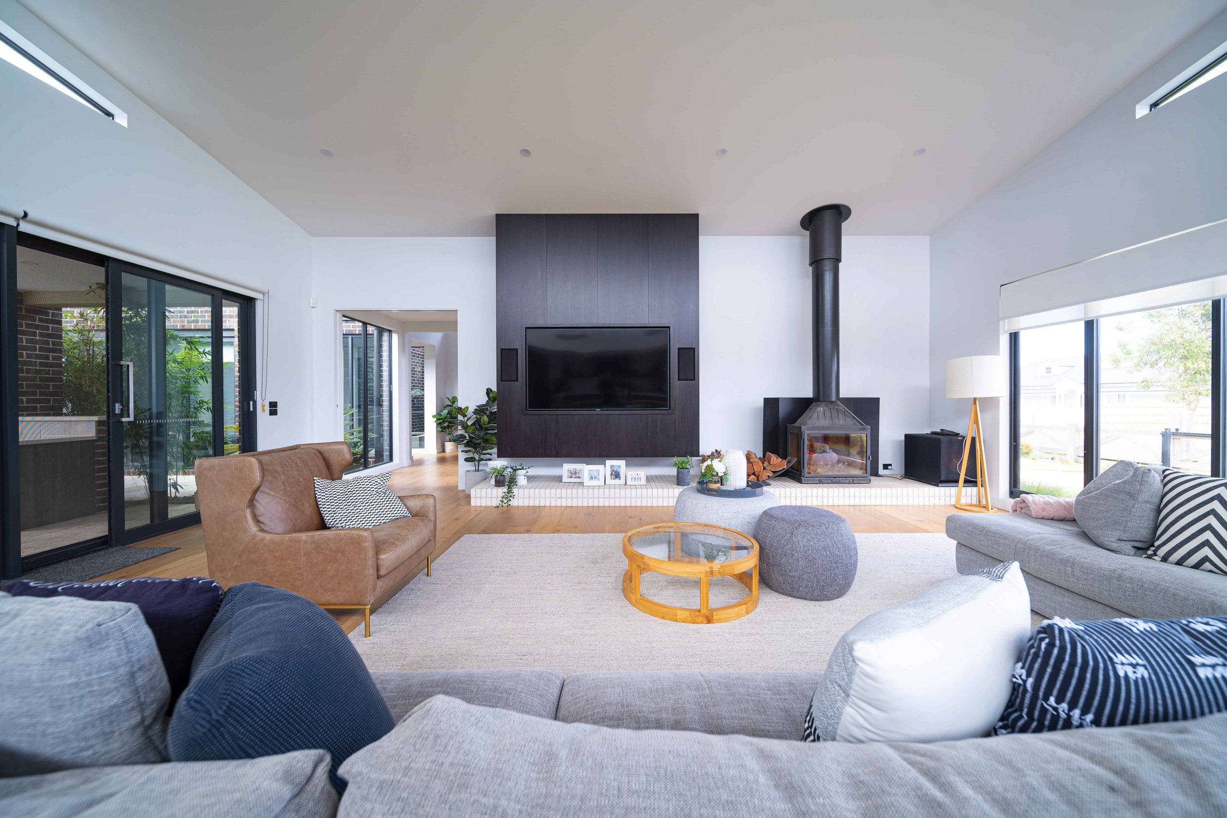 Modern living room with a raked ceiling. Features a black wood log fireplace, a wall-mounted TV in a dark wood panel cabinet, and large windows with blinds.