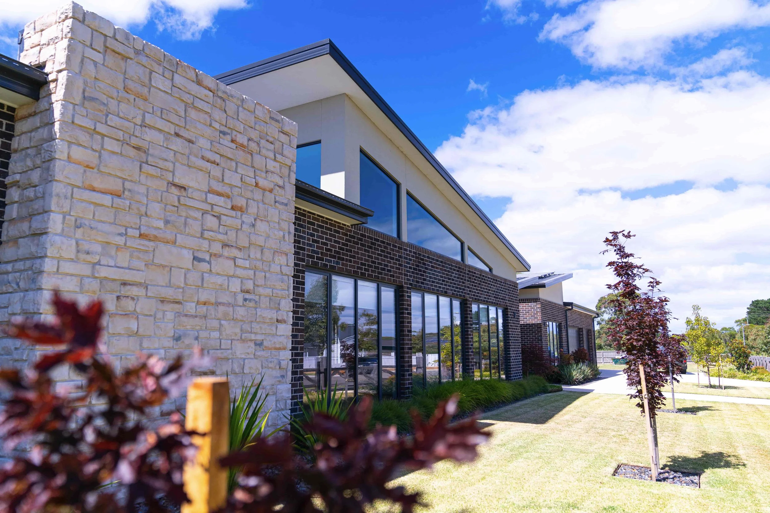 Modern residential building with brick and stone exterior walls, large glass windows, and a landscaped yard with a pathway, trees, and plants under a blue sky with clouds.