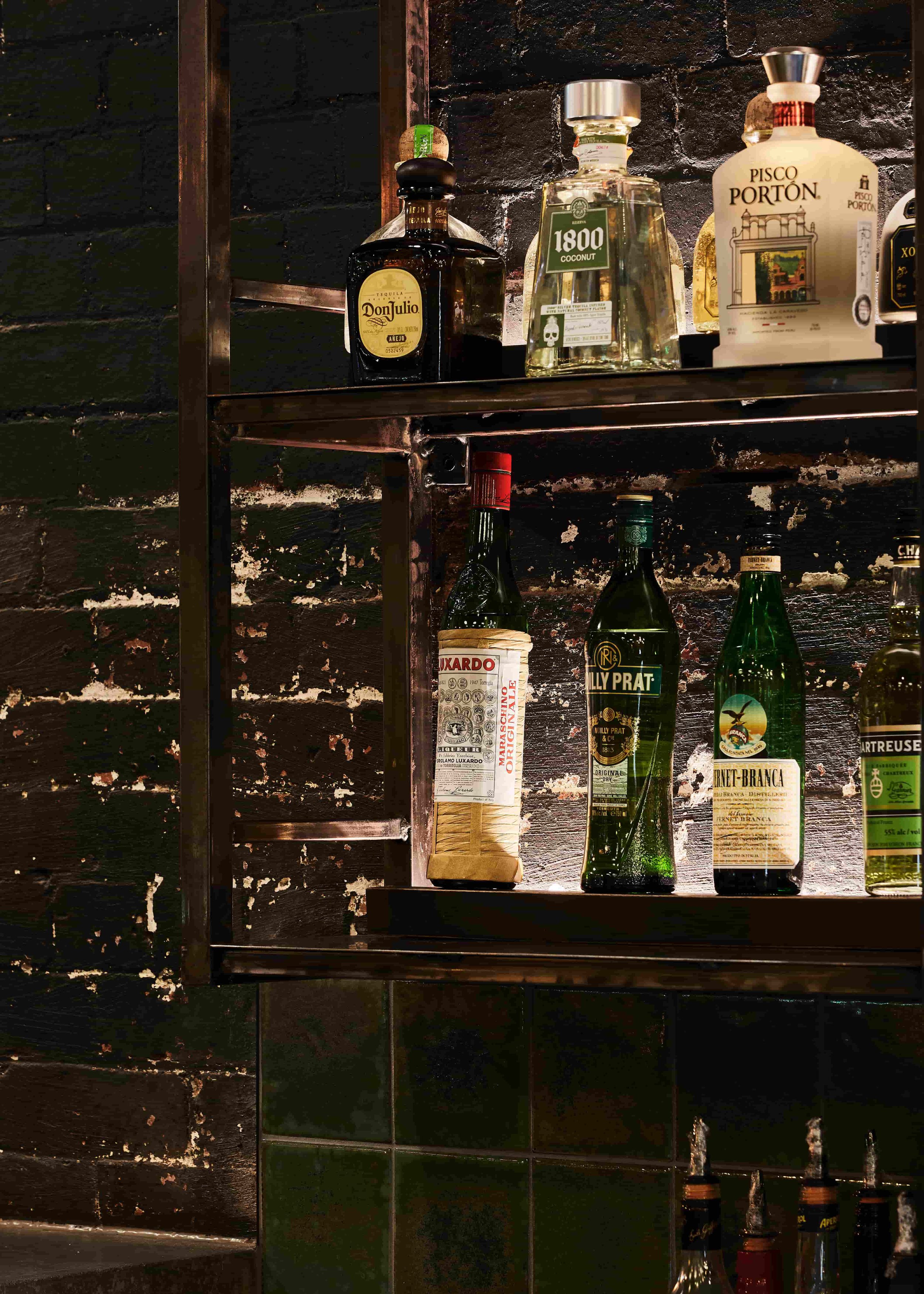 Shelves with bottles of alcohol and condiments against a black, textured brick wall.