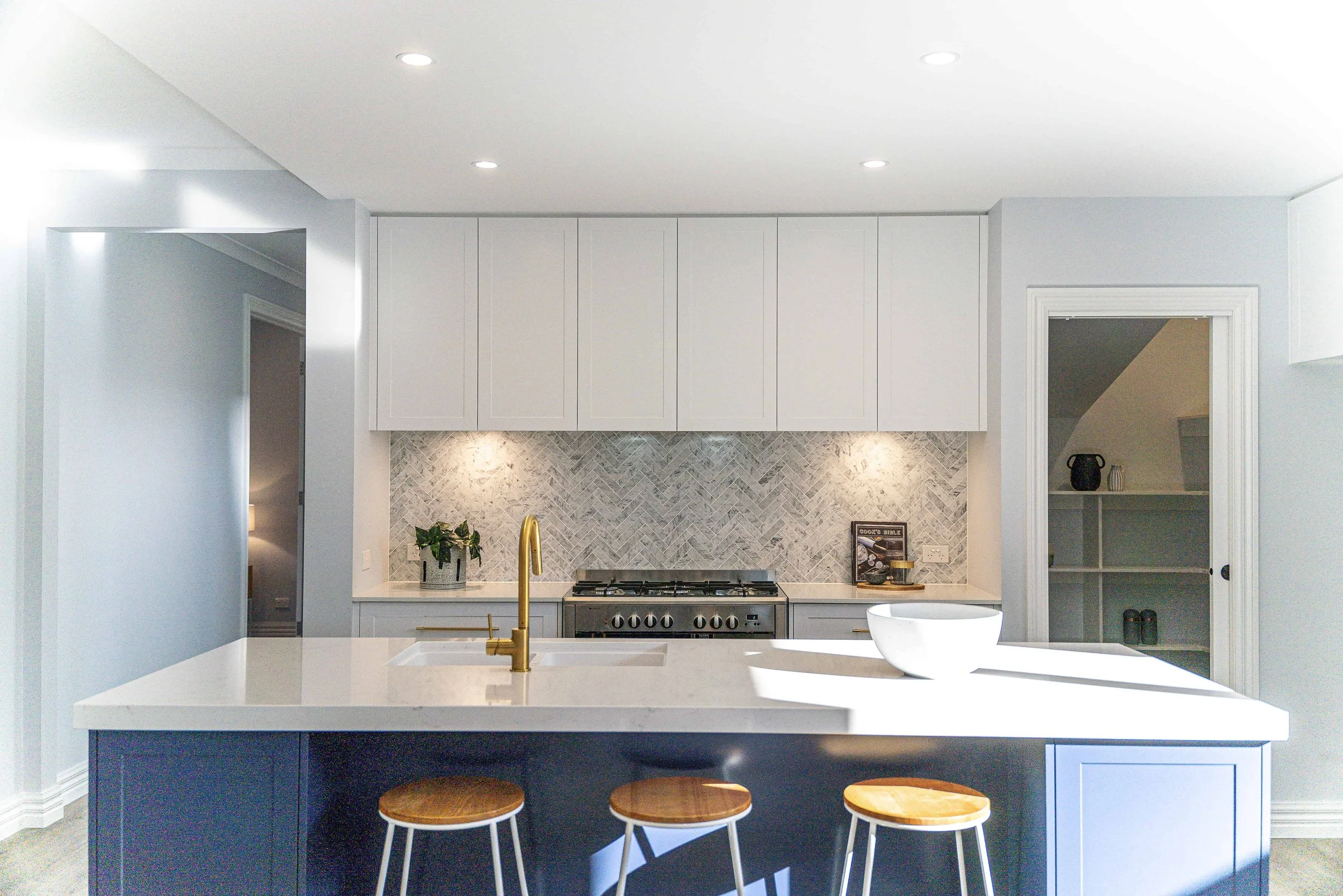 Modern kitchen with a blue island, white stone benchtops, and bar stools with wooden seats. Ash coloured wood flooring and grey walls. The island has routed door panels.