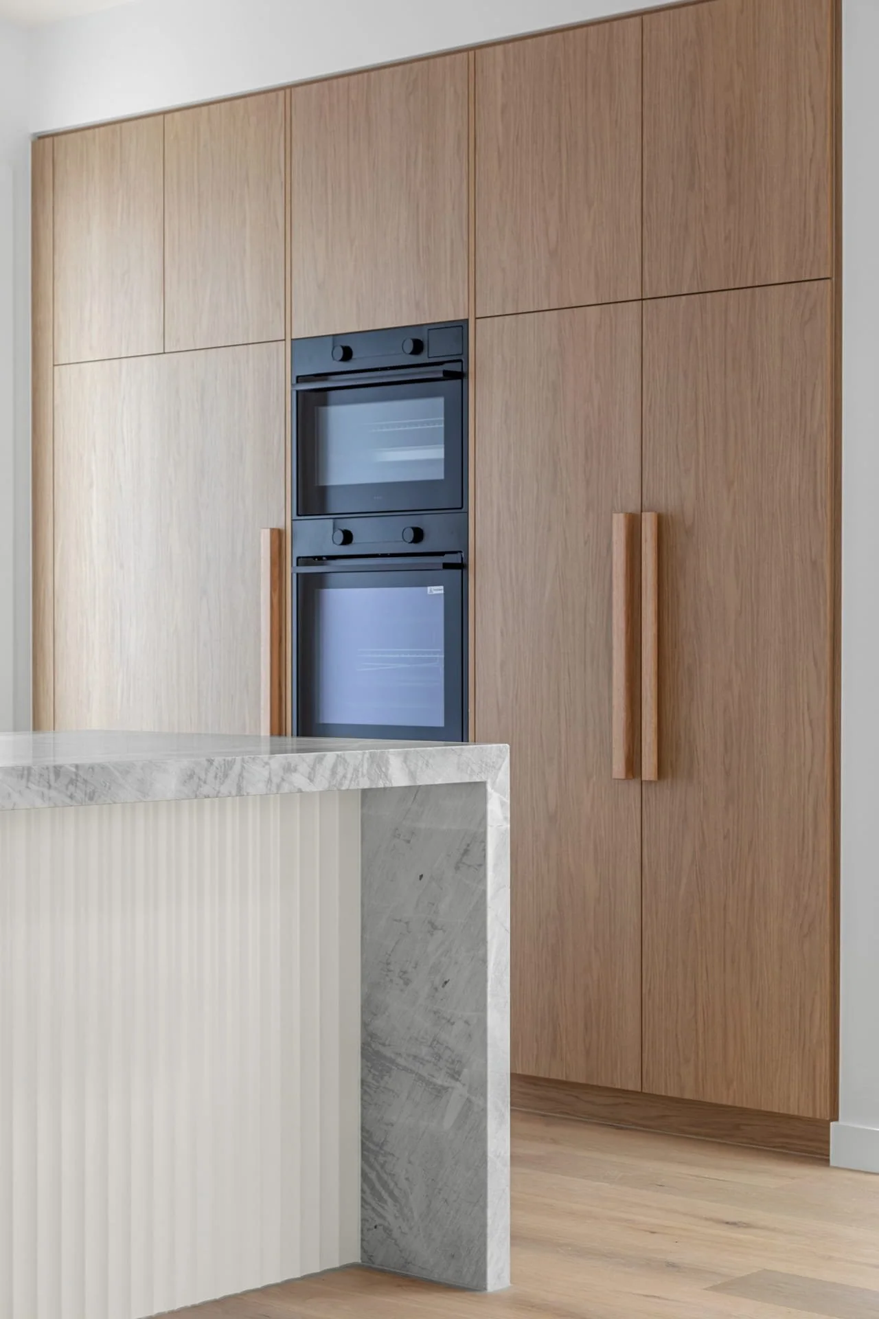 Modern kitchen with tall timber cabinets to the ceiling, built-in black ovens, a grey stone island benchtop with an overhang and waterfall finish, and scalloped white island panelling.