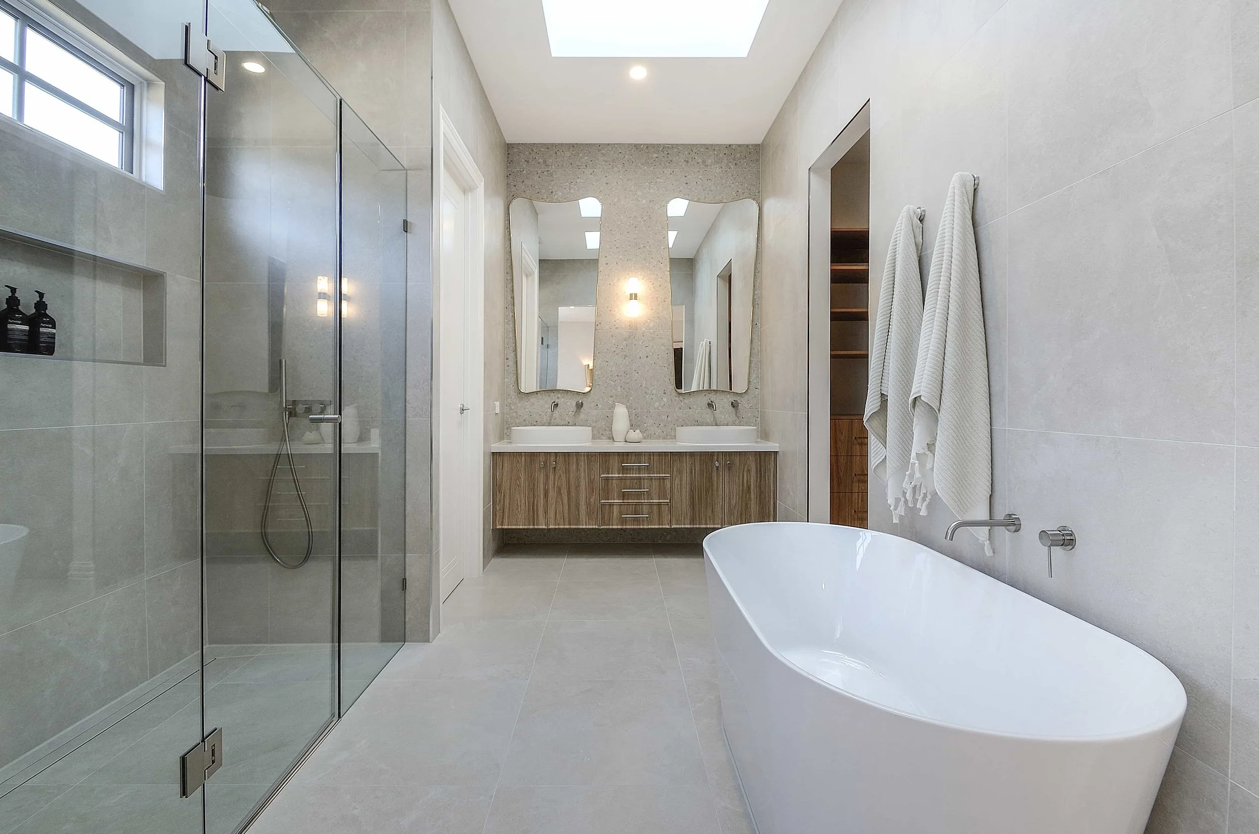Modern ensuite with a walk-in shower, double sink vanity with feature gold framed mirrors, a freestanding bathtub, beige tiled walls and floor, white towels hanging on the wall, and a small window and skylight.