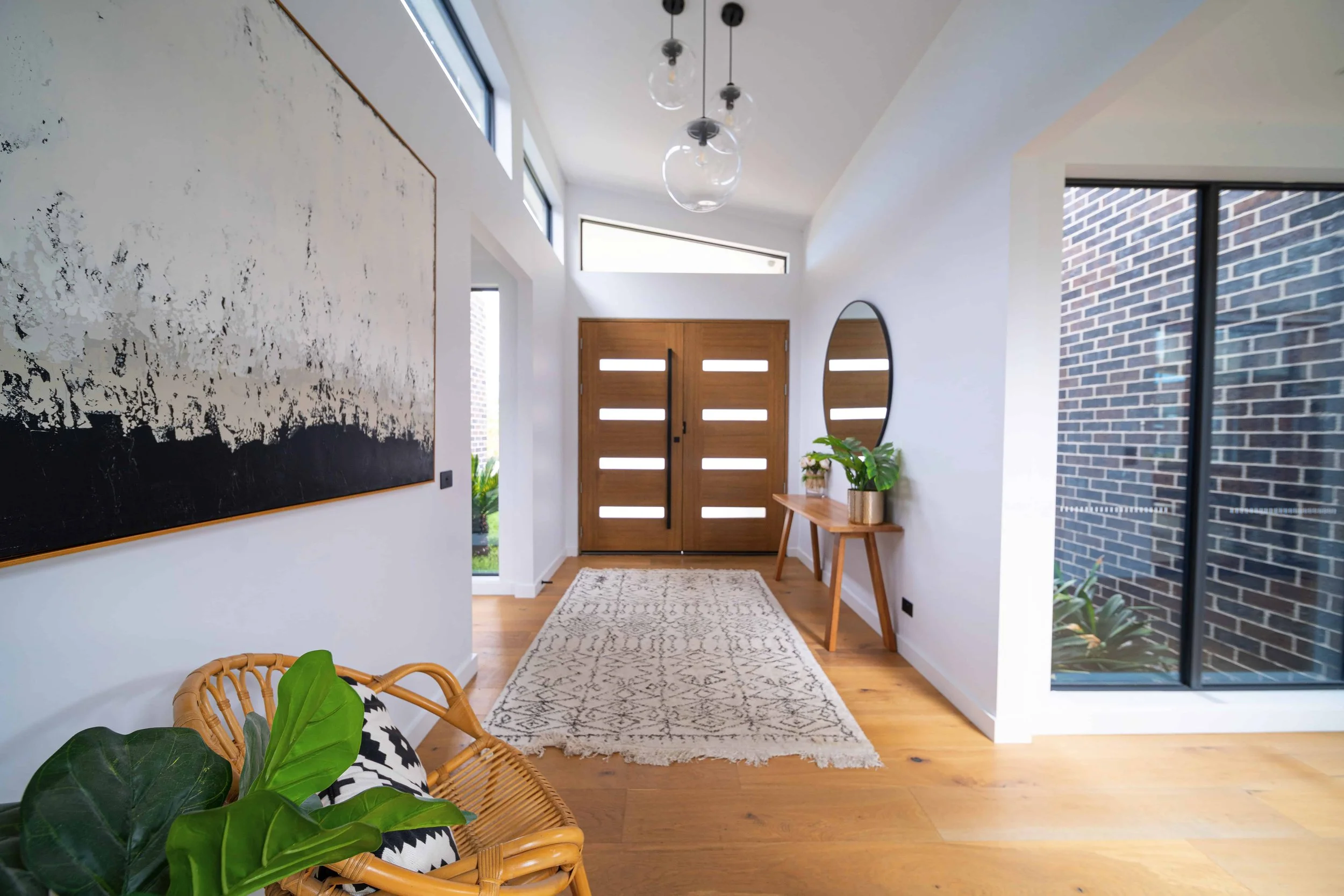 Modern entryway with wooden double doors, a large mirror, a small wooden table with a potted plant, and a white rug. Interior features include white walls, wooden flooring, a brick wall, and pendant lights with glass globes. Natural light comes throu