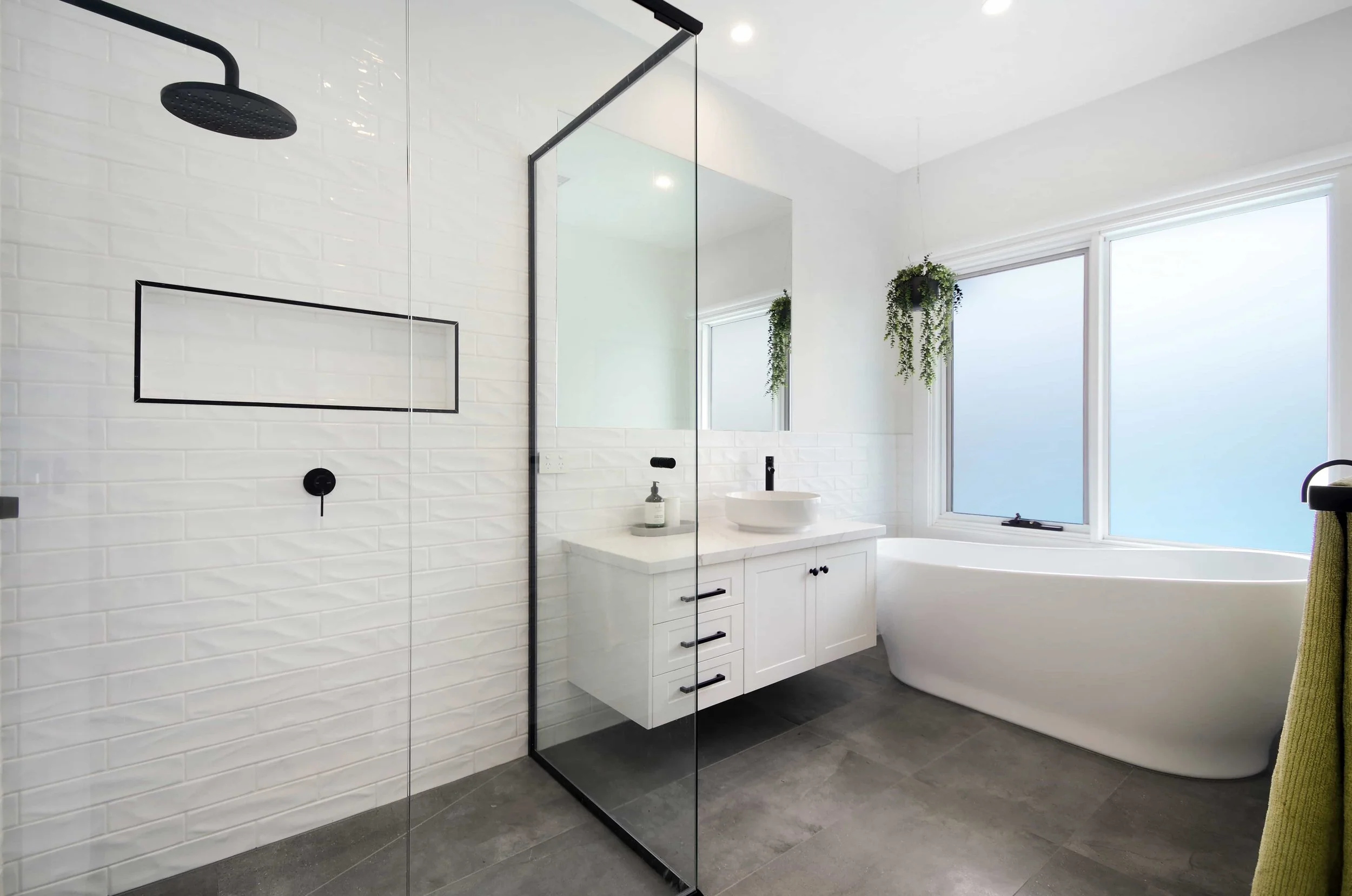 Modern bathroom with a glass enclosed shower, a floating white vanity with black handles and a round vessel basin above counter, a large mirror, a freestanding bathtub near a window with frosted glass, and hanging green plants.
