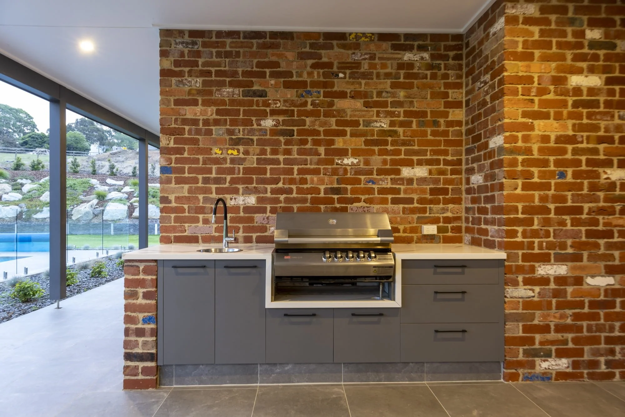 Outdoor barbecue area under an alfresco, with modern grey cabinets, a stainless steel bbq, next to a backyard with a pool and landscaped garden.