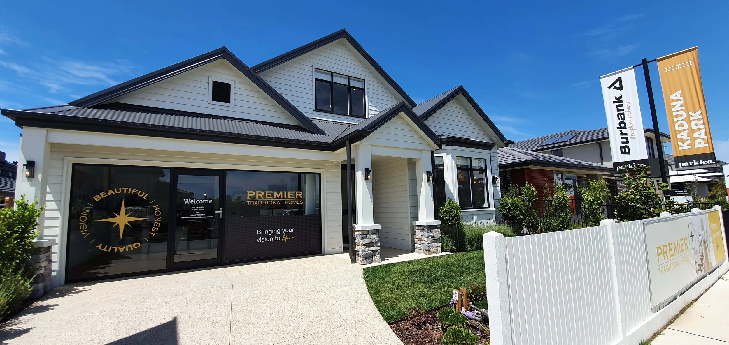A modern Hamptons style house with a white weatherboard exterior and monument roofing. There is a feature porch with stone clad piers and thick square columns. It is a display home in the Burbank area with the Premier Traditional Homes branding.
