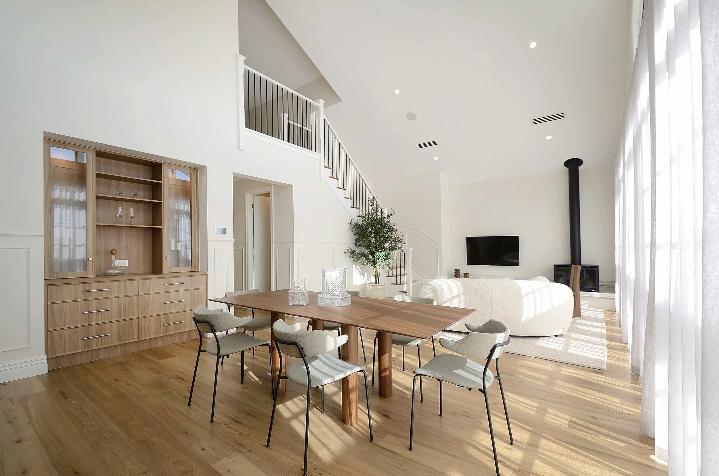 Open living and dining room with cathedral ceiling, light timber floors, large windows with sheer curtains, white walls, staircase, wall-mounted TV, wood log fireplace, and dining table with six chairs.