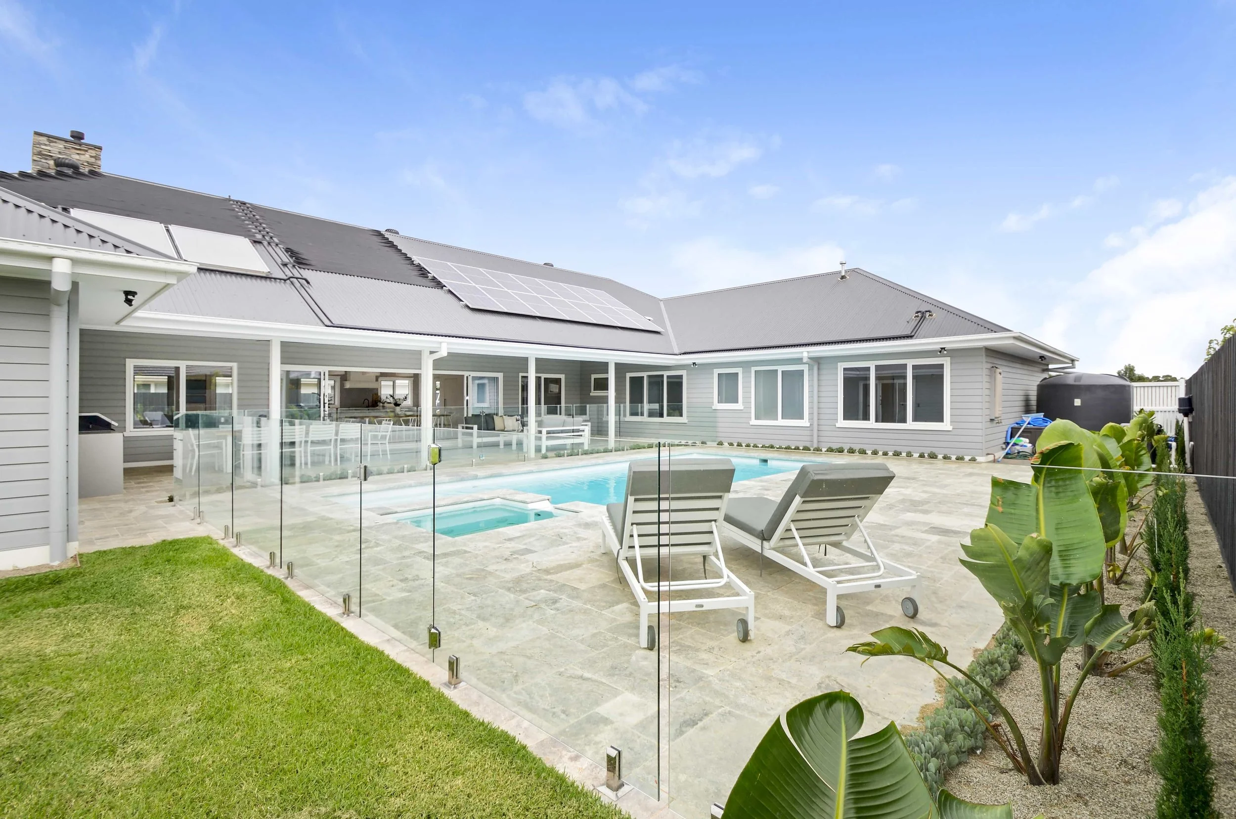 A backyard with a swimming pool, lounge chairs, and a house with solar panels on the colorbond roof. The house is in grey weatherboard with feature white trims.
