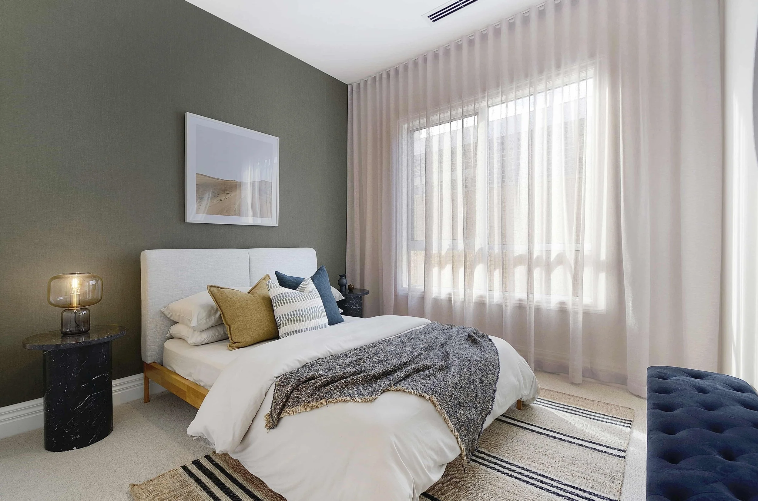 Modern bedroom with a white bed, multiple pillows, and a gray throw blanket. Nightstand with a glass lamp, dark bedside table, large window with sheer curtains, and minimalist wall art. There is a green olive feature textured wall behind the bed.