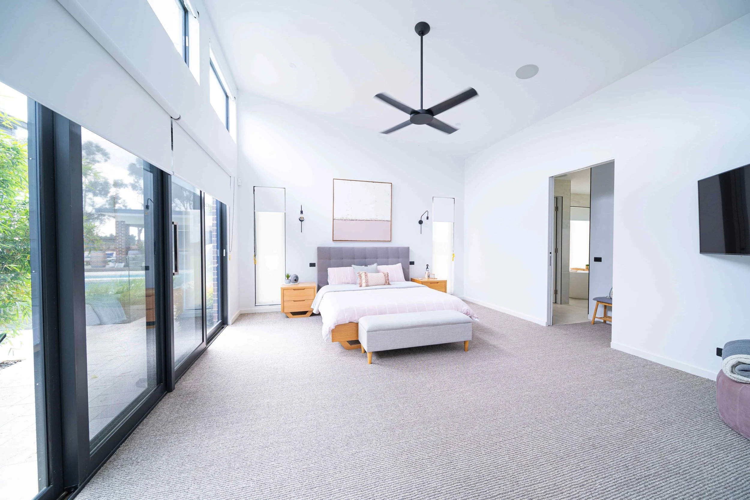 A modern master suite with a large bed, light wood bedsides, and a grey upholstered bench. The room has tall windows and doors with black frames, white walls, and a ceiling fan. The feature ceiling is raked.