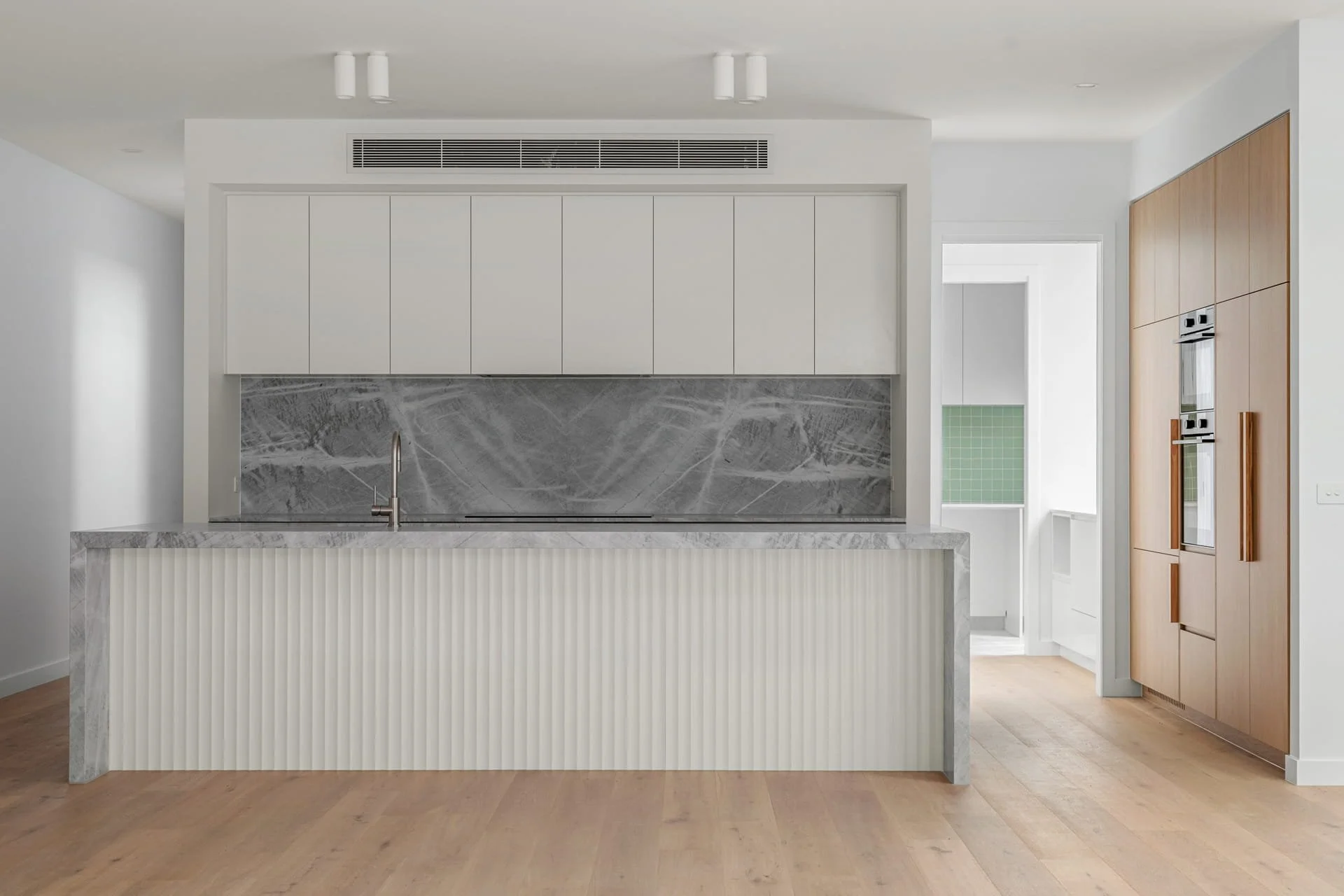 Modern kitchen with a stone island, waterfall finish, white upper cabinets, and a built-in oven in timber cabinets, with natural light coming through a window. The back panel of the island is scalloped.