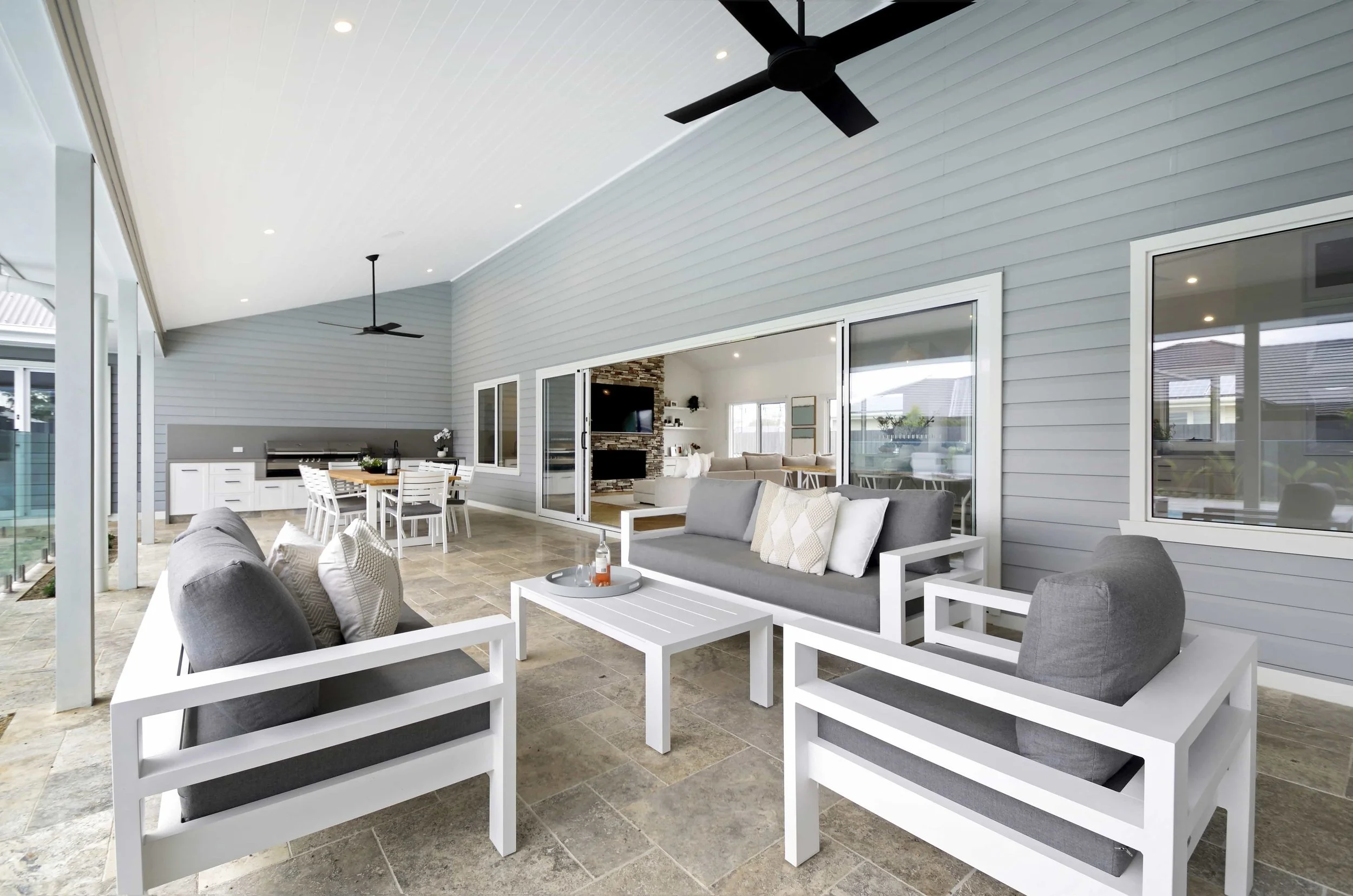 An alfresco area with modern seating, ceiling fans, and a large sliding door leading to the open living space beyond. The is an outdoor barbecue area on the far wall.