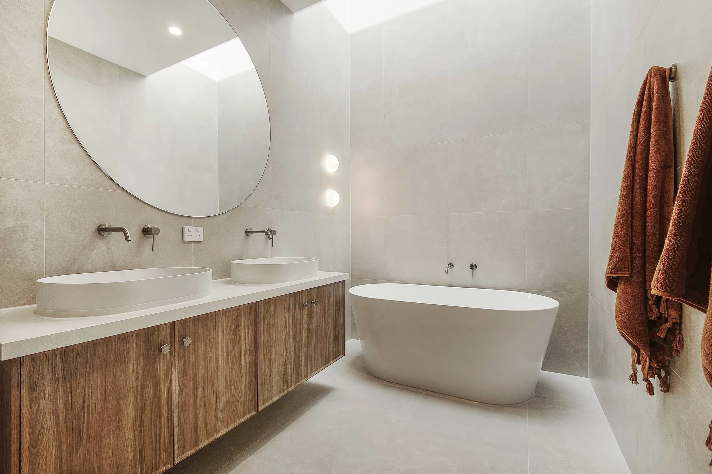 Modern bathroom with a large round mirror, a double sink vanity with timber cabinetry, a freestanding bathtub, beige tiled walls, and a brown towel hanging on a rack. There is a skylight over the bathtub.