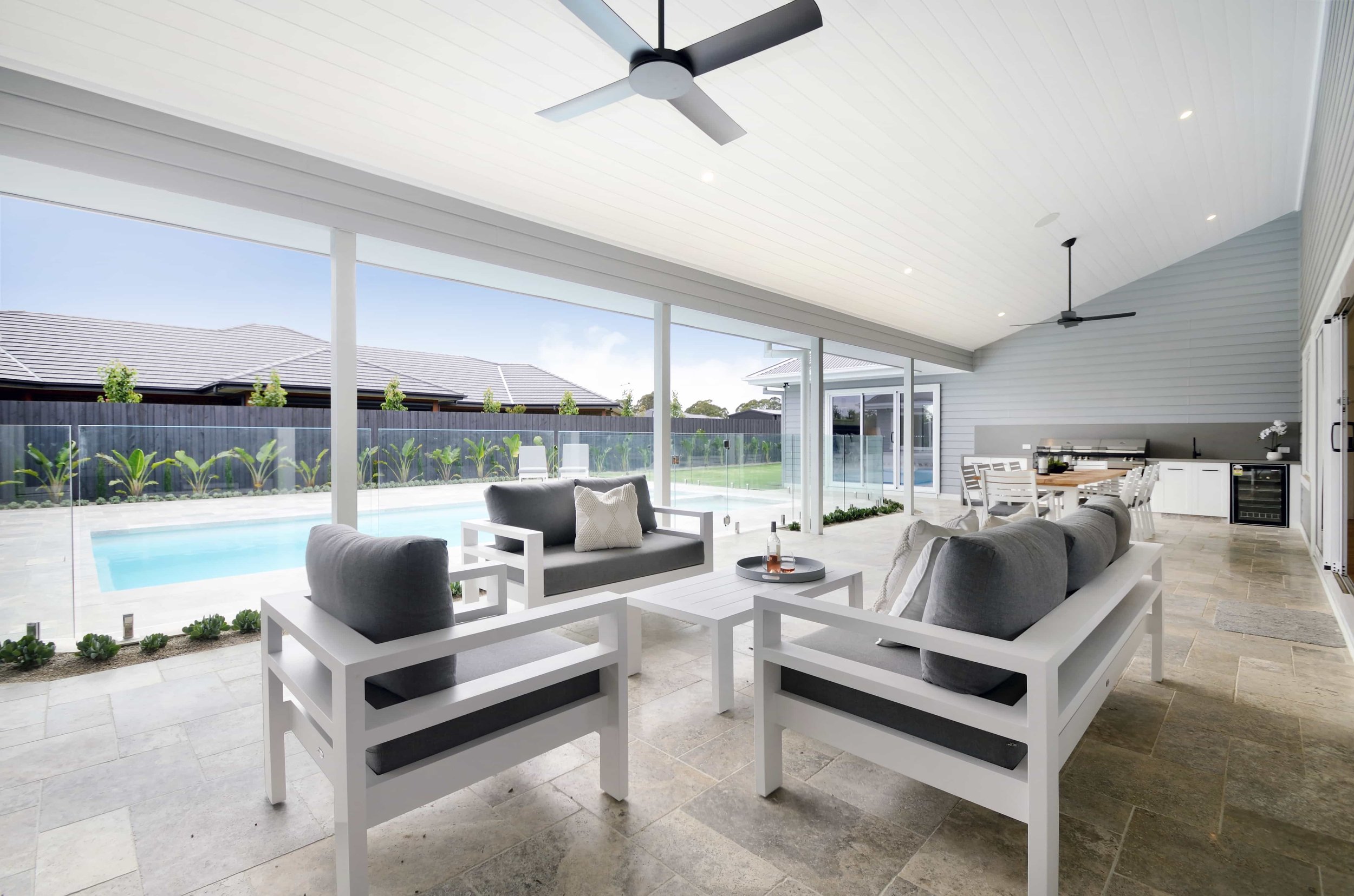 An alfresco area with modern seating, ceiling fans, and a view of a backyard pool and garden. The ceiling above the alfresco is raked and lined with VJ board.