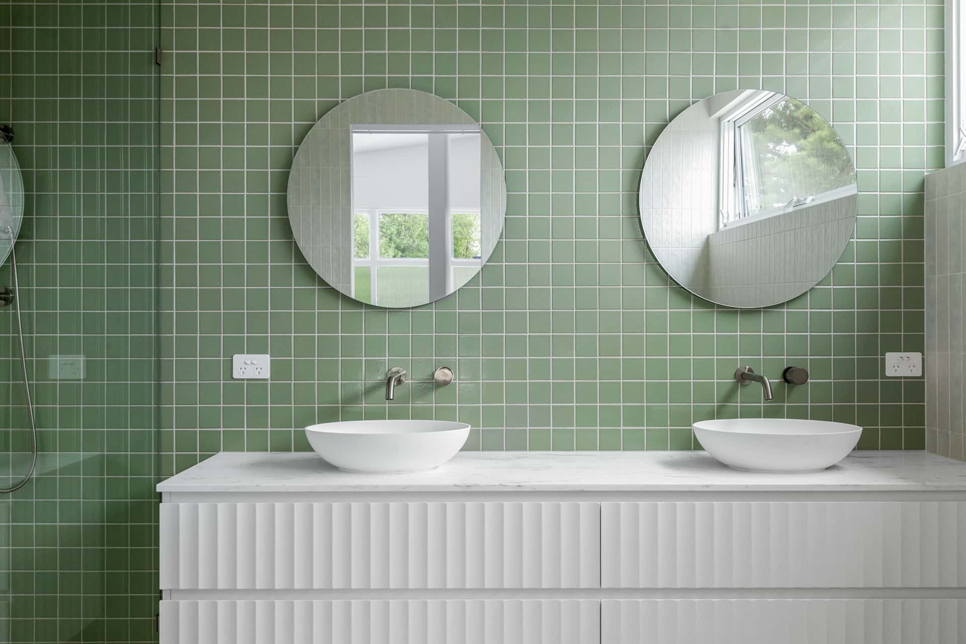 A modern ensuite with two round mirrors above two oval above bench basins, green mosaic tiled wall, and scalloped vanity doors.
