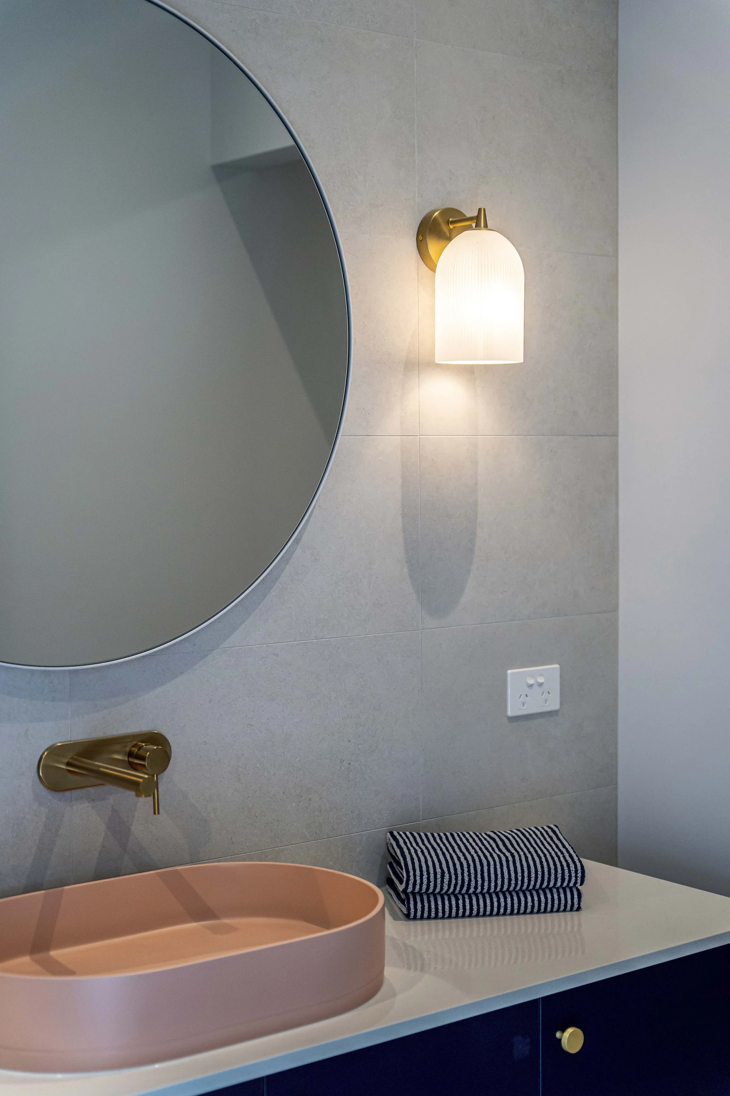 Bathroom with a large round mirror, a pink sink, a gold faucet, a wall-mounted light fixture, a striped towel, and a power outlet on a grey tiled wall.