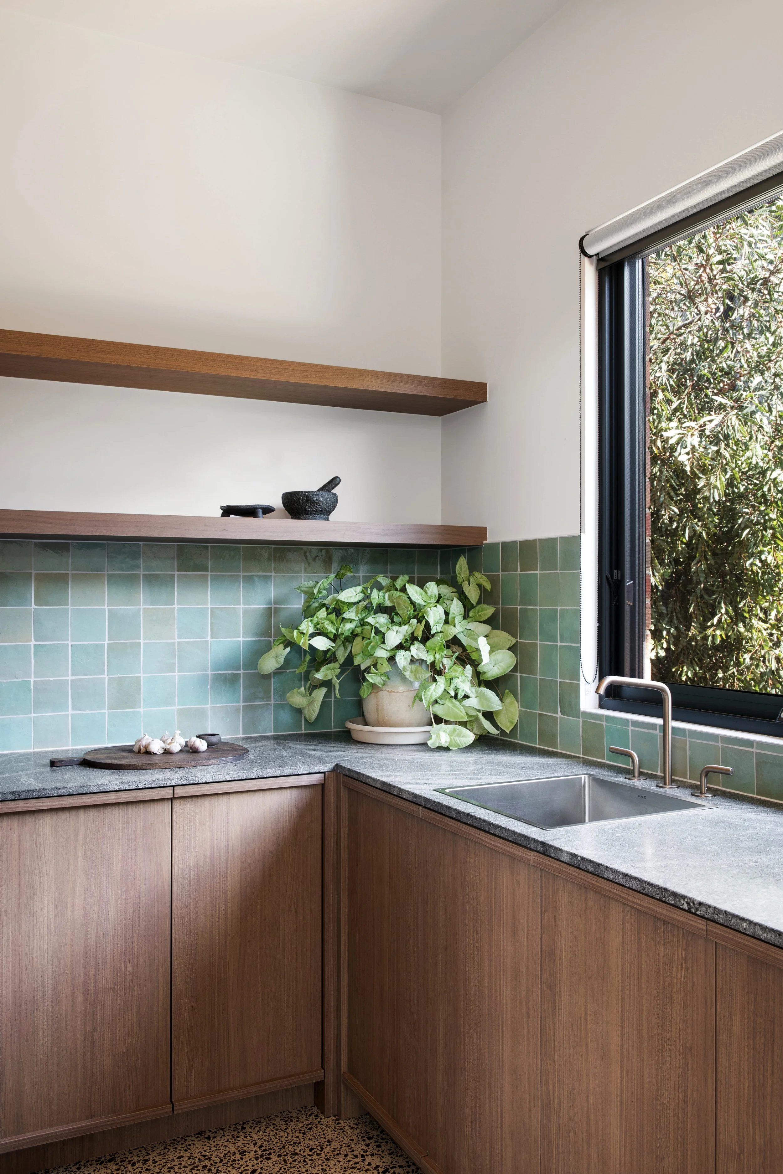 A kitchen with timber cabinets, a grey stone benchtop, green tiled splashback, and a window with a view of greenery.