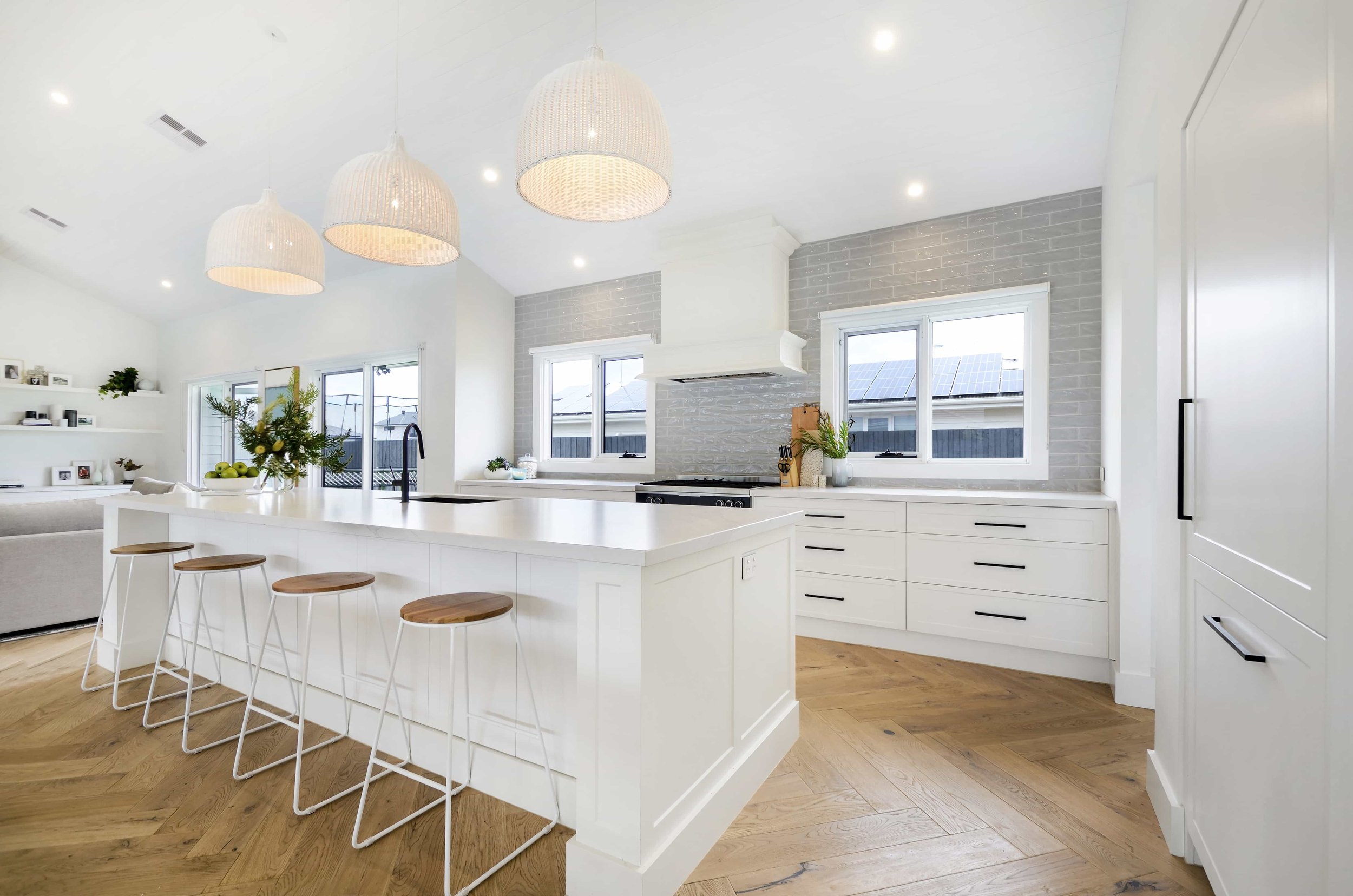 Modern white kitchen with large island, feature pendant lights, glossy grey subway tile splashback, two windows, and timber flooring in a herringbone pattern.