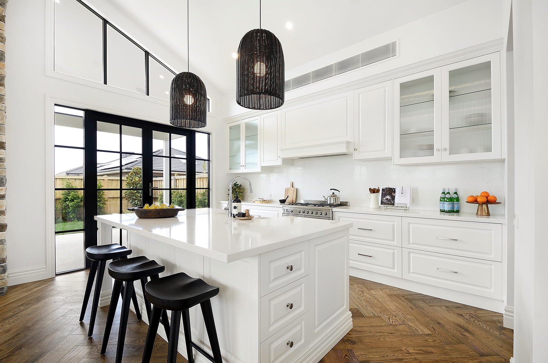 Modern white kitchen with routed white door panels, black wicker pendant lights over the island bench, black bar stools, and large glass French doors leading outside.