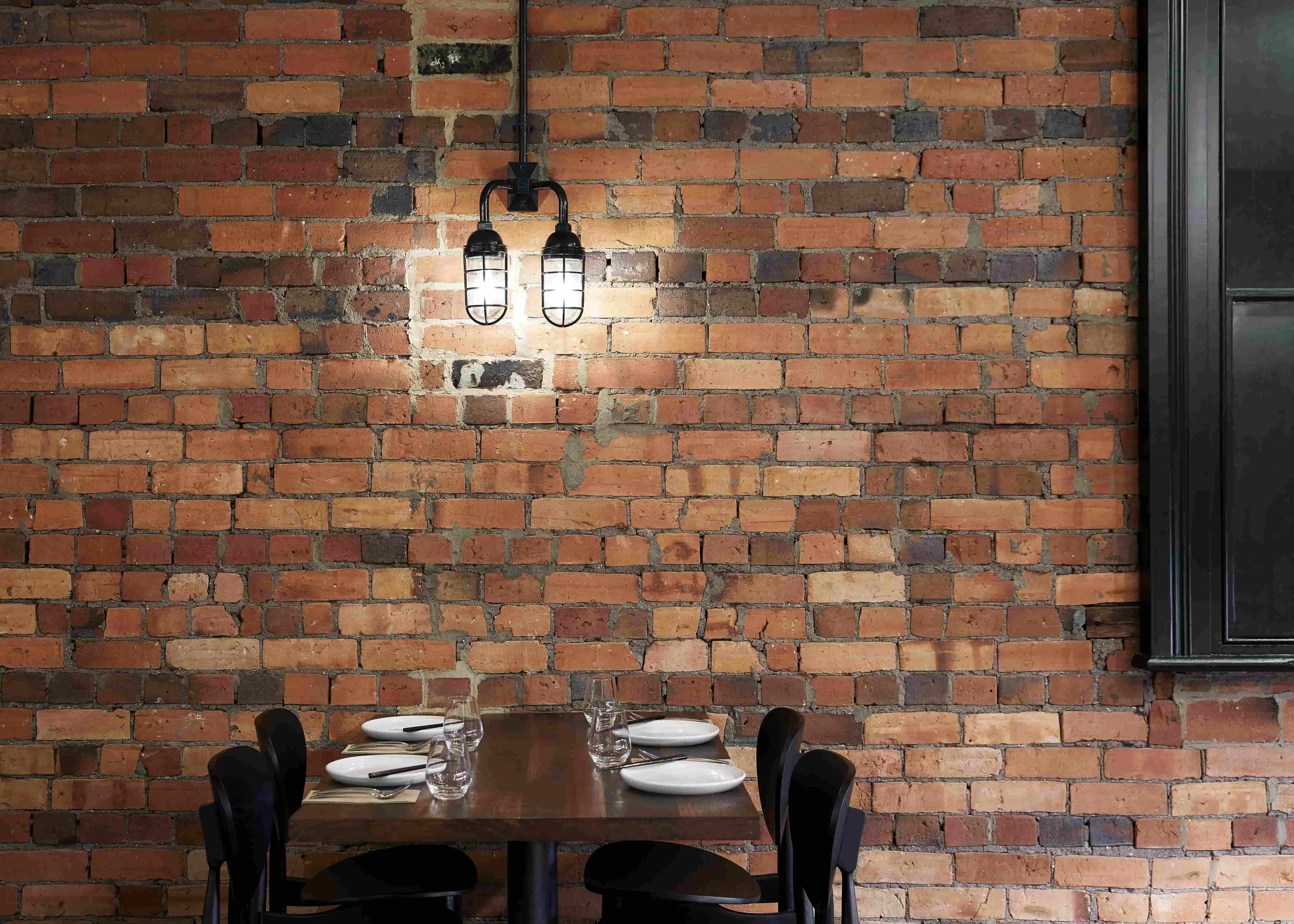 A dining table set for four, with white plates, glasses, and utensils, against a brick wall with industrial-style lighting above.