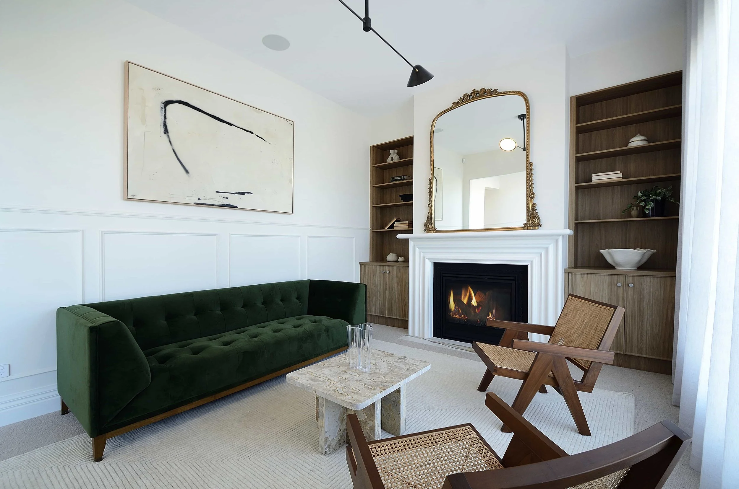 Formal living room featuring a green velvet sofa, white walls with decorative wainscoting, a fireplace and traditional mantel with ornate gold-framed mirror above, and built in timber bookshelves.