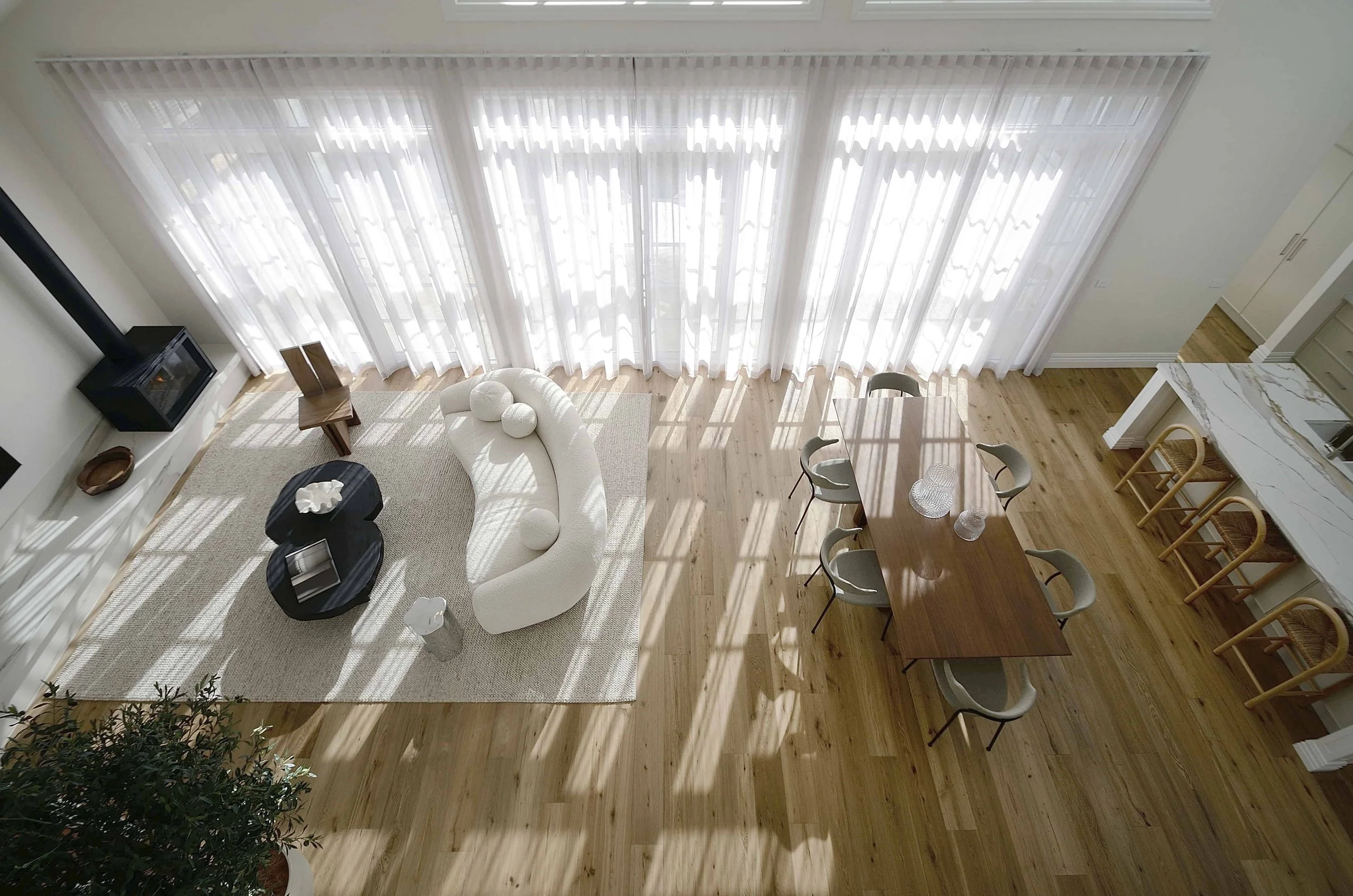 Spacious open living room with large windows covered by sheer white curtains, a white couch, a black coffee table, a TV on the wall, a dining table with six chairs, and timber flooring, with sunlight casting shadows through the curtains.