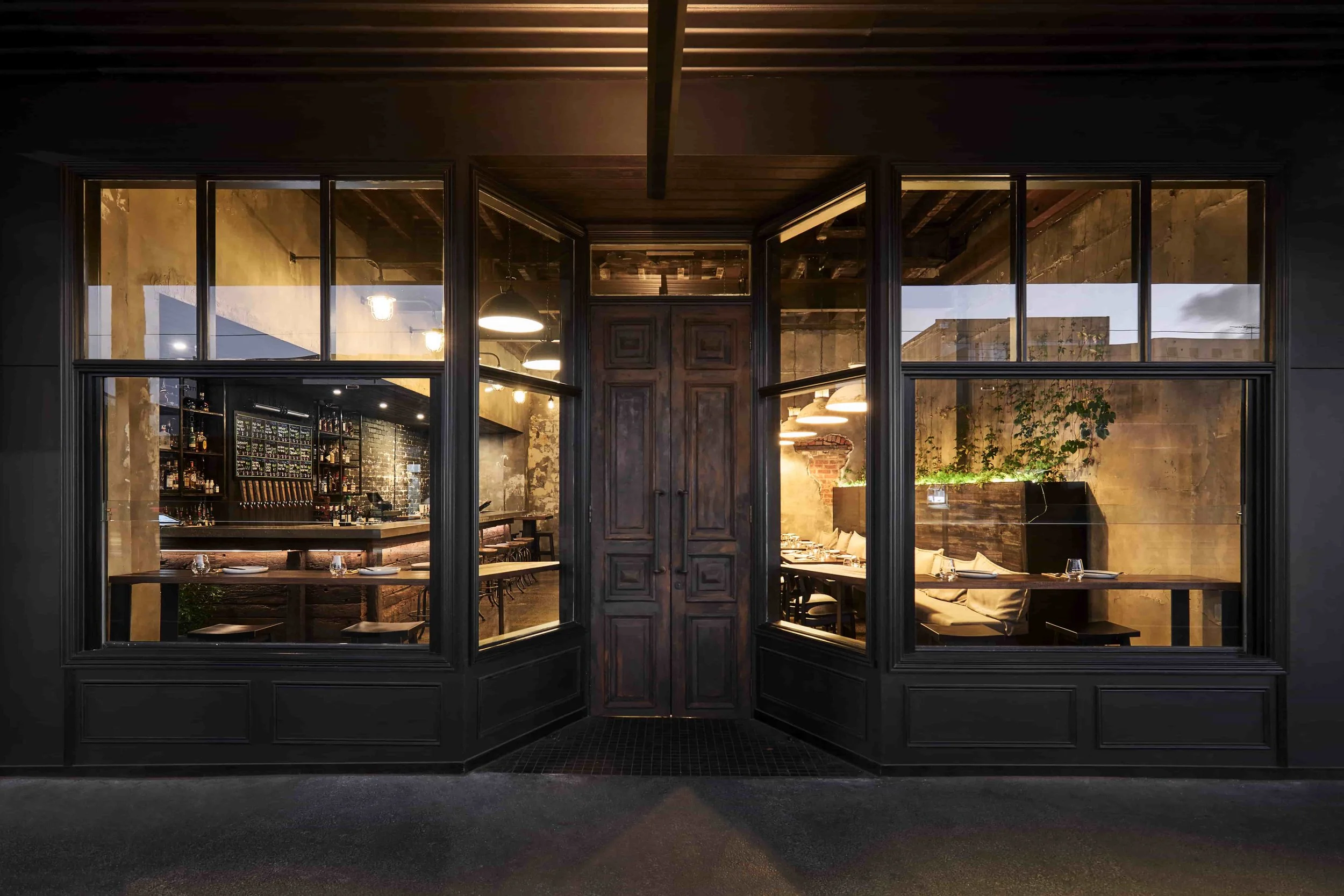 View through a large open shopfront window into a warmly lit dining area of a restaurant with exposed brick and concrete walls, ceiling lights, and a long dining table with chairs.