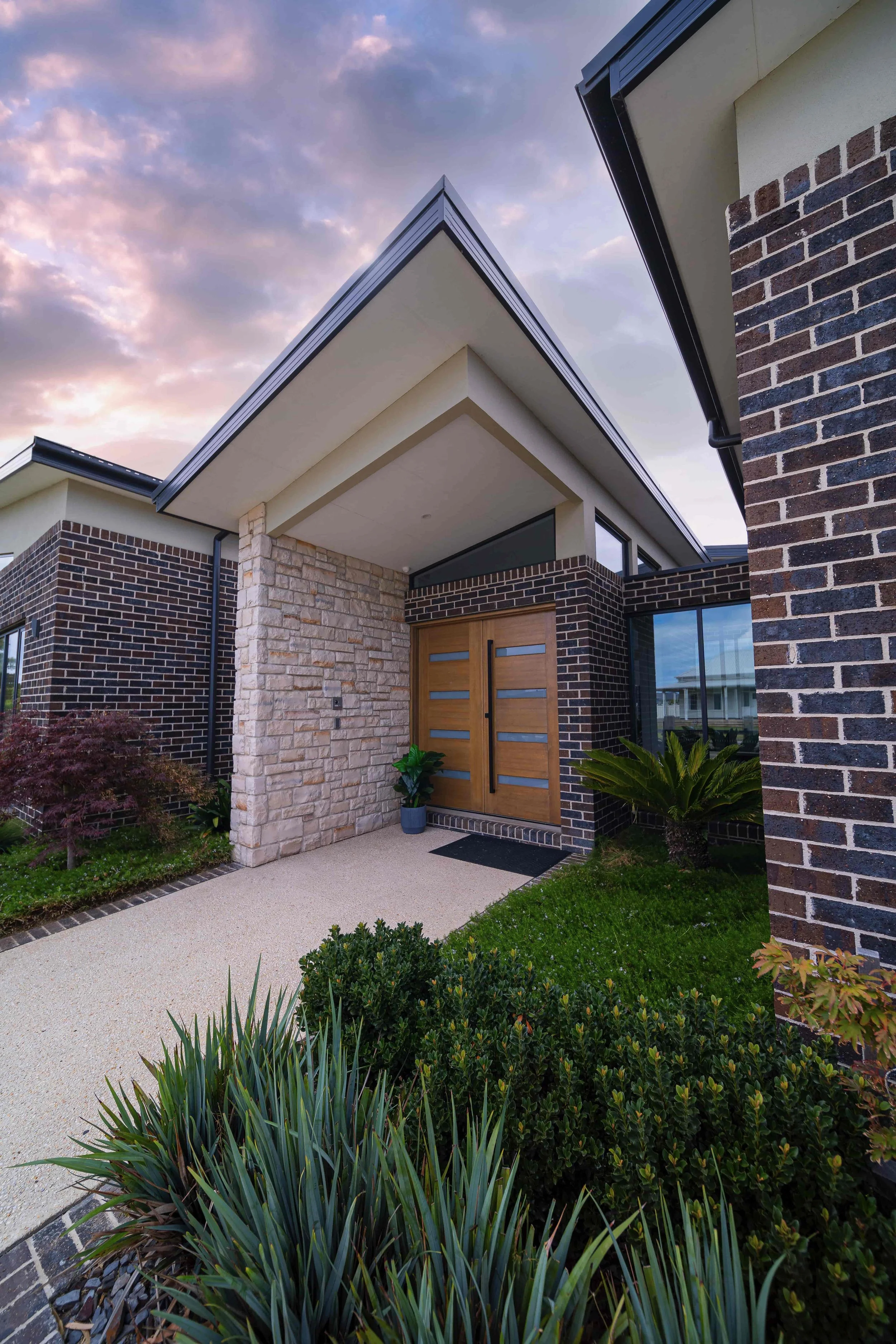 Modern house entrance with dark brick and light stone exterior, wooden door, landscaped front yard with various plants, and a cloudy sky at sunset.
