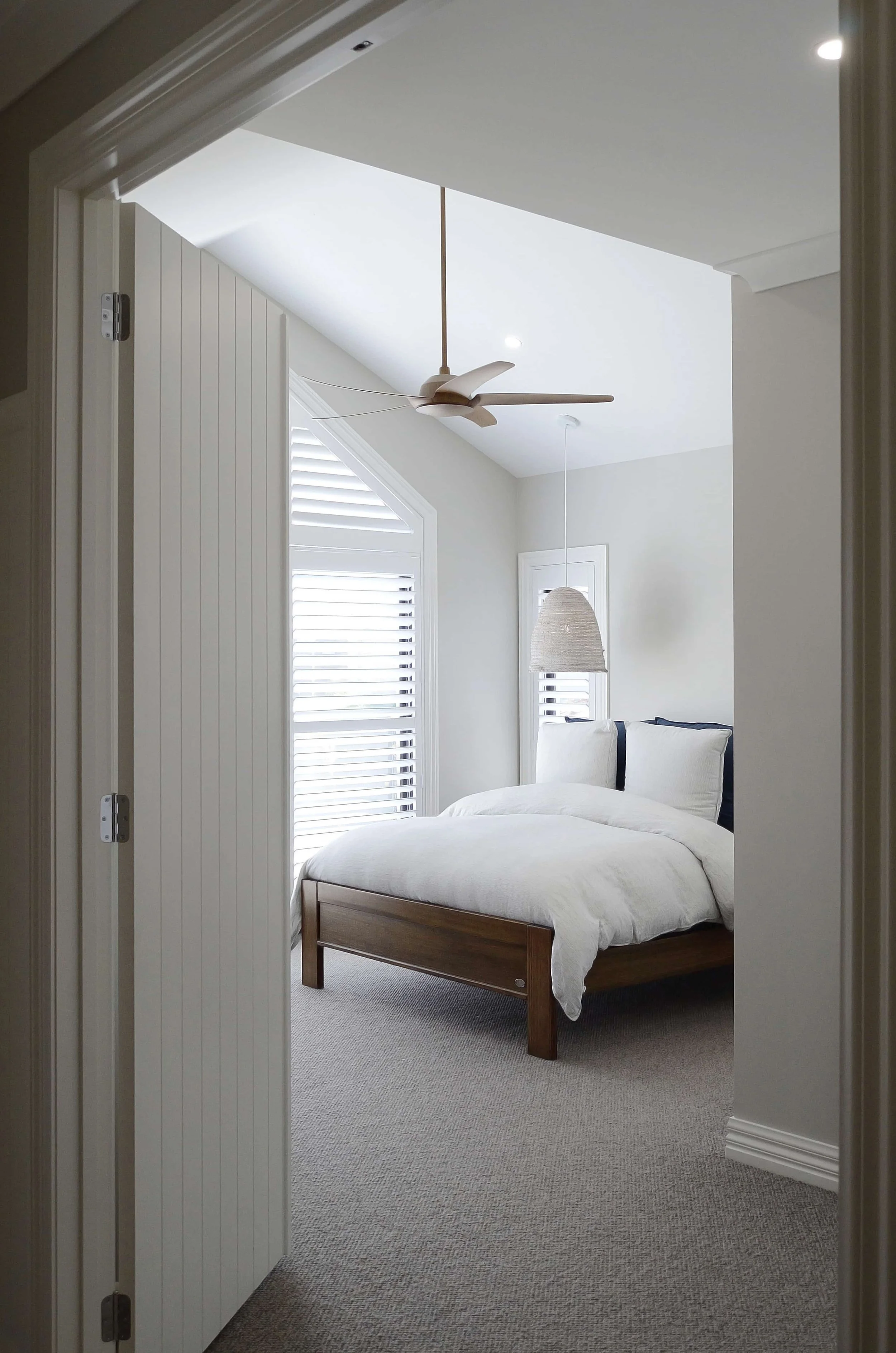 A bedroom with a view through a doorway showing a bed with white bedding, light gray walls, a ceiling fan, and window blinds.