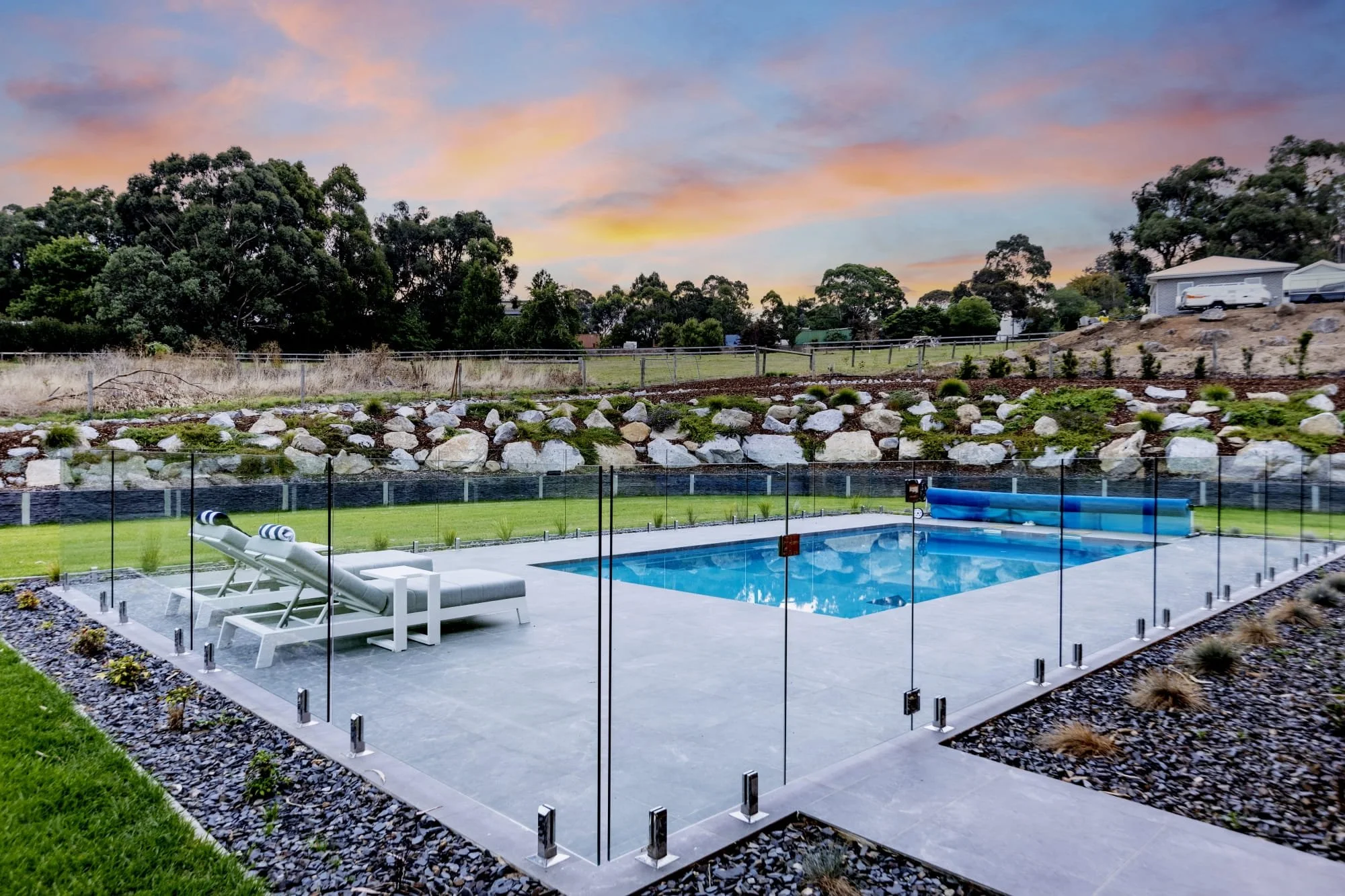 A backyard with a fenced-in swimming pool and lounge chairs, bordered by rocks and plants, with a hillside and trees in the background during sunset.