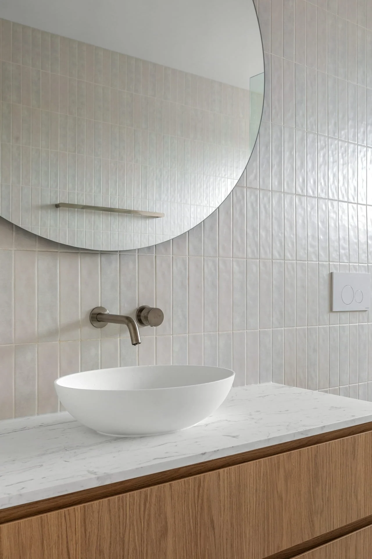 Modern bathroom with a white above bench basin on a white marble benchtop, a large round mirror, beige vertical subway tiled wall, and a wooden vanity with a natural finish.