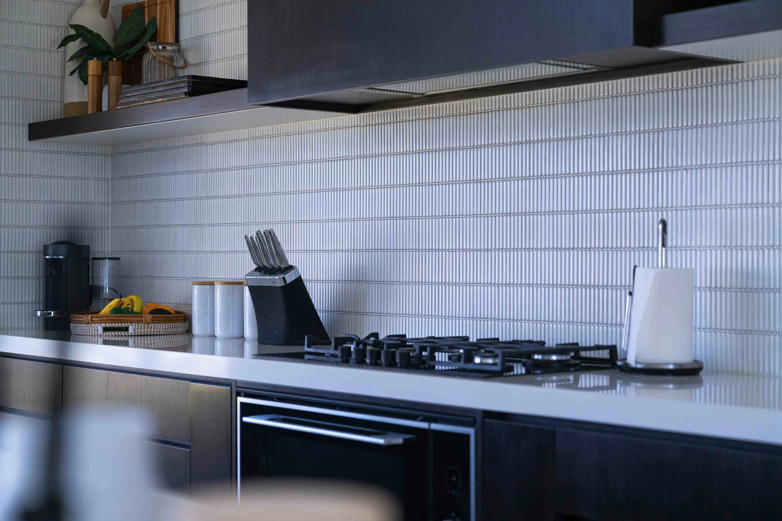 Modern kitchen with a white benchtop, black stove and cook top, and a white finger tile splashback. There's a black concealed undermount rangehood above and matching floating shelves on either side.