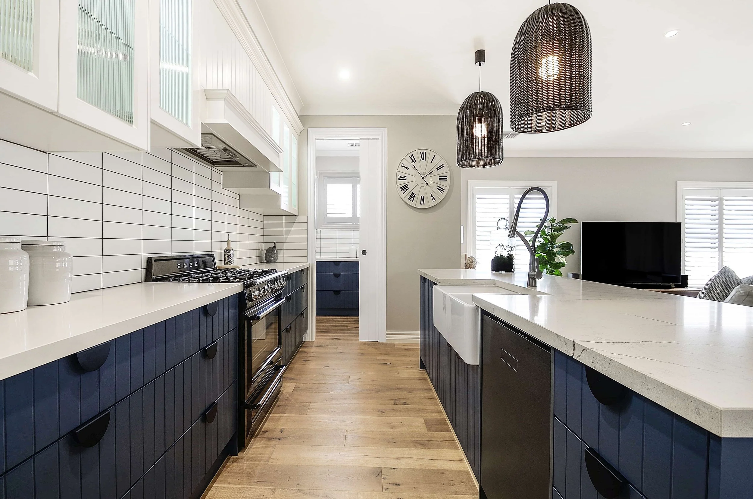 Modern kitchen with a blue island, white stone benchtops, black rattan pendant lights above the island, and bar stools with wooden seats. Light-coloured wood flooring, grey walls, and large windows. The cabinetry has vertical routing in the panels.