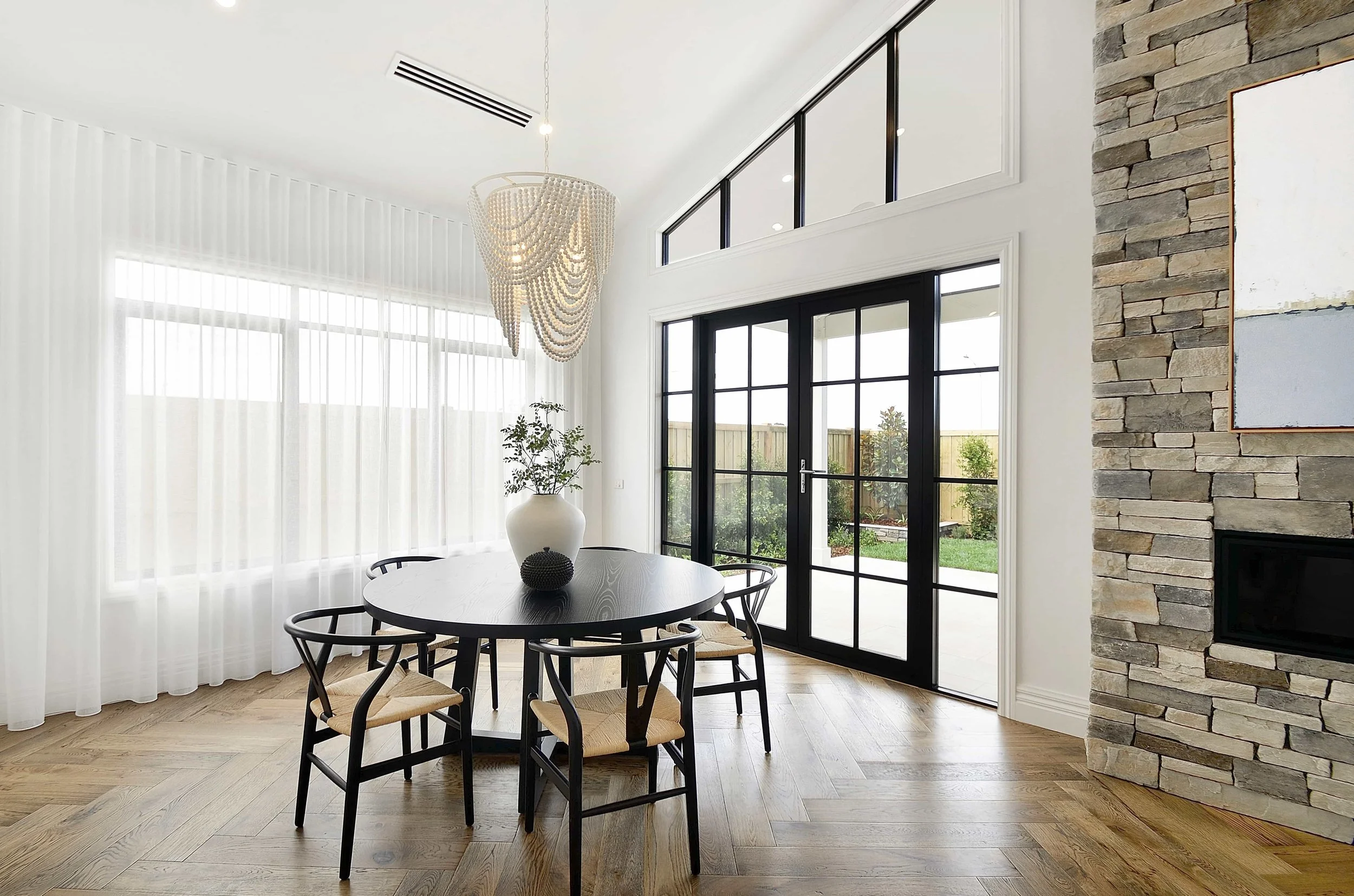 Modern dining area with a round wooden table, five black chairs with woven seats, large colonial windows with white curtains, a decorative chandelier, a potted plant, and a stone fireplace wall.