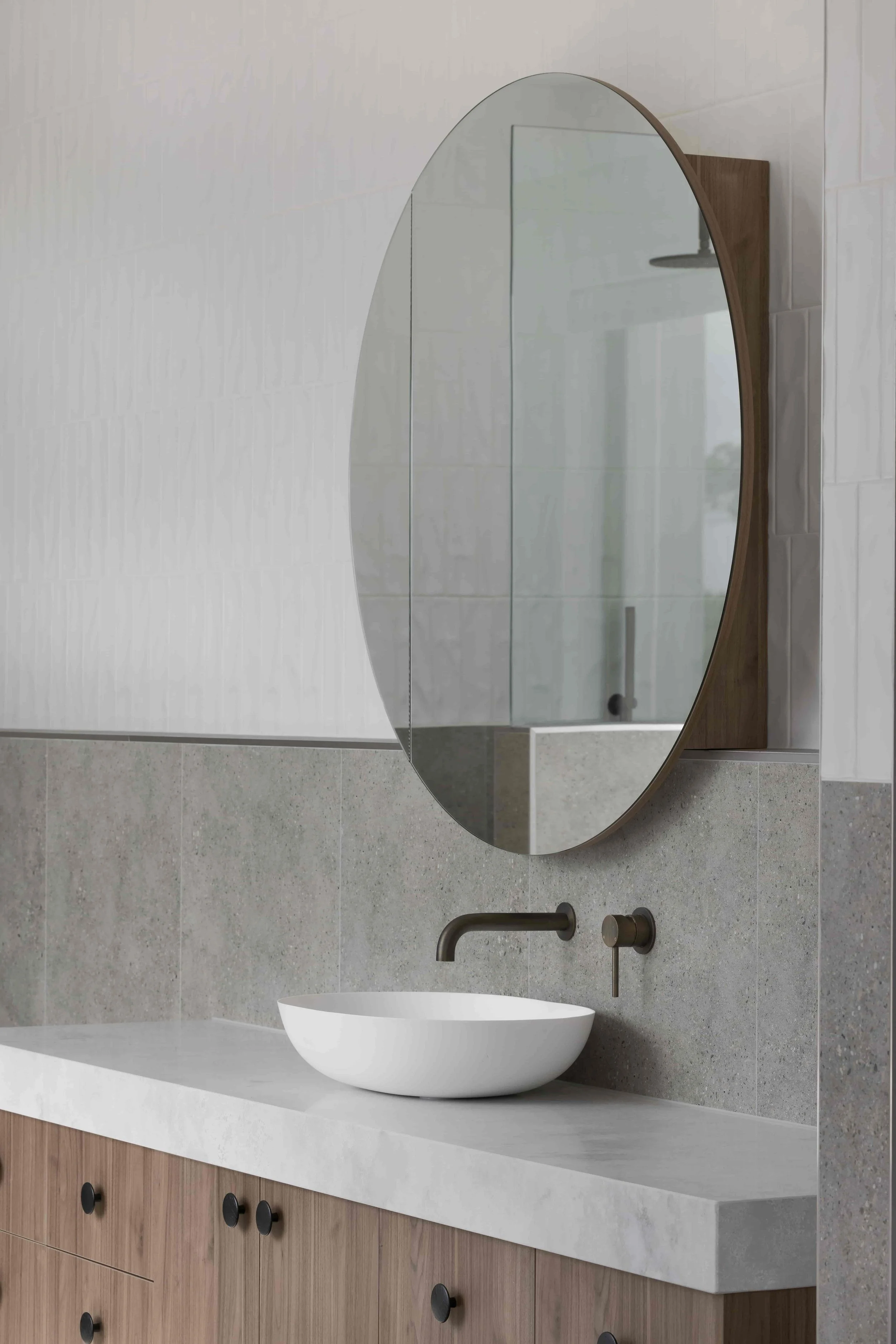 Modern bathroom vanity with a white above bench basin, thick grey benchtop, speckled dark grey wall tiles, timber cabinet with round black handles, round wall mirror, and wall-mounted bronze faucet.
