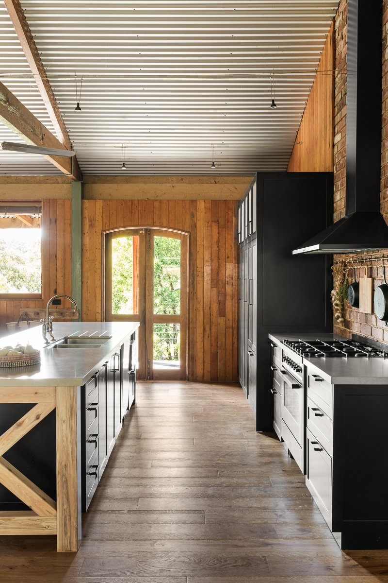 Modern rustic kitchen with timber lined walls, natural timber flooring, black cabinetry, copper accessories, and a brick accent wall.
