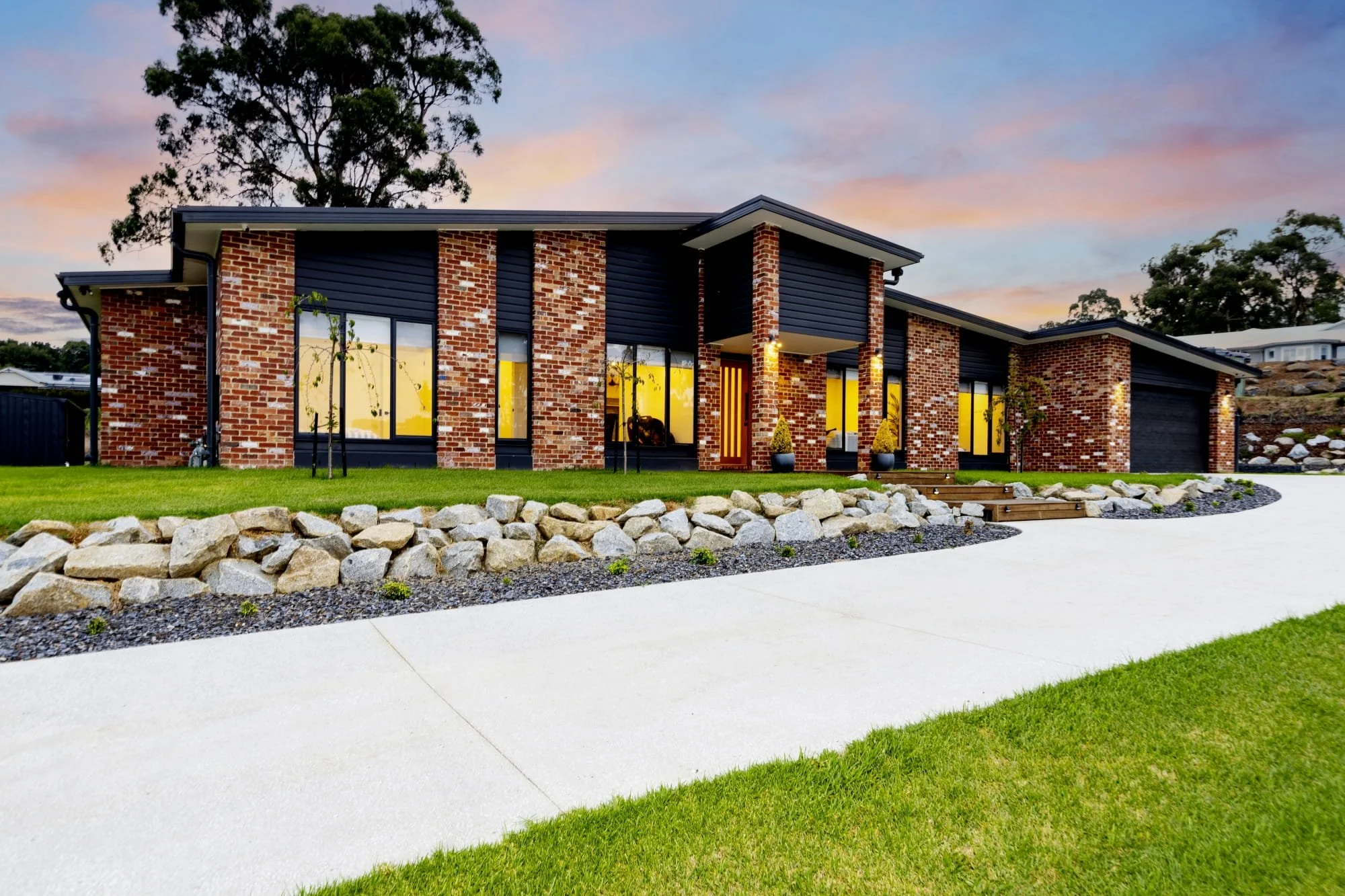 Modern brick and black weatherboard house with large windows, front porch, and landscaped yard during sunset.
