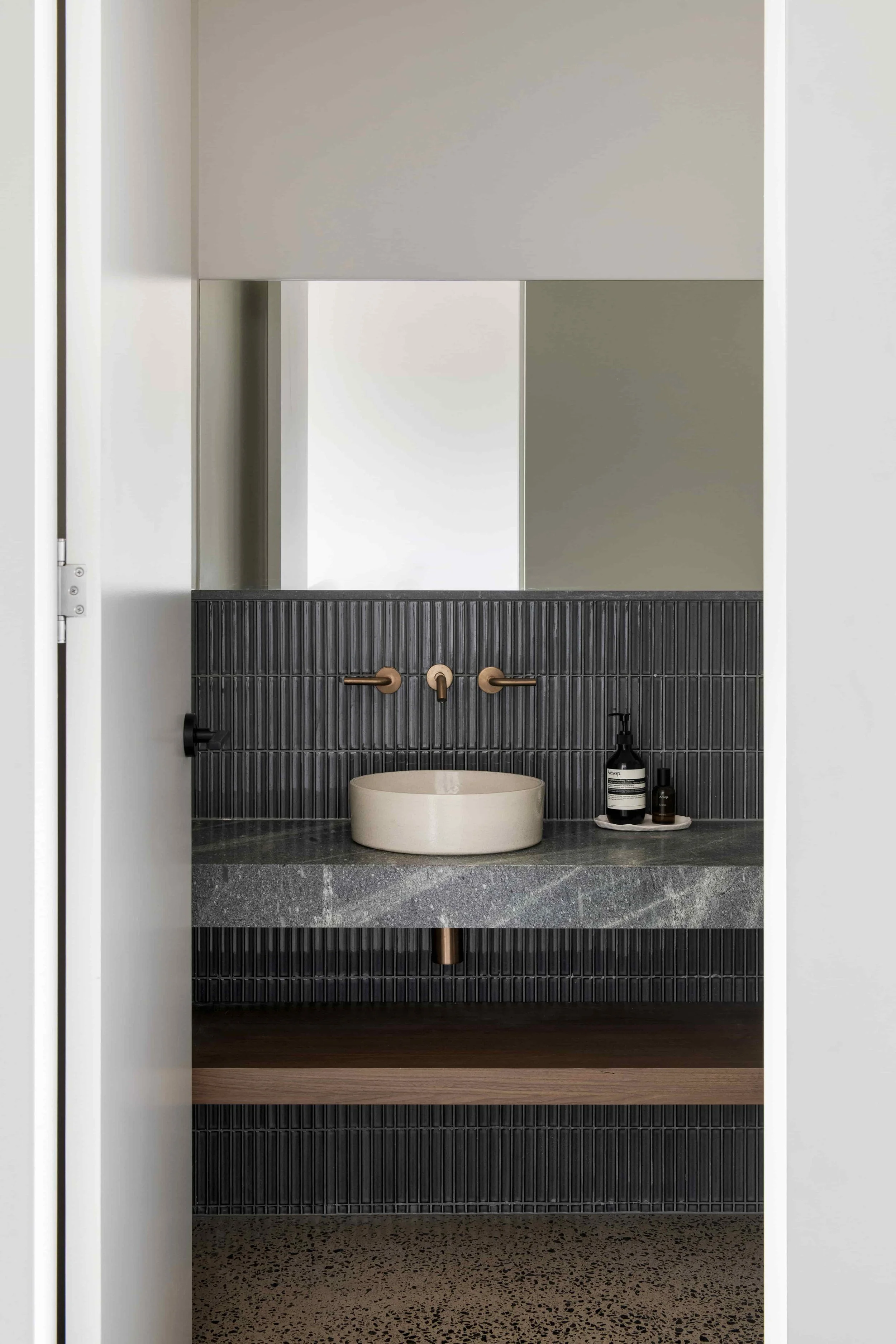Bathroom with black tiled accent wall, tan vessel sink, black soap dispenser, and bronze faucet, with dark terrazzo flooring, seen through a doorway.