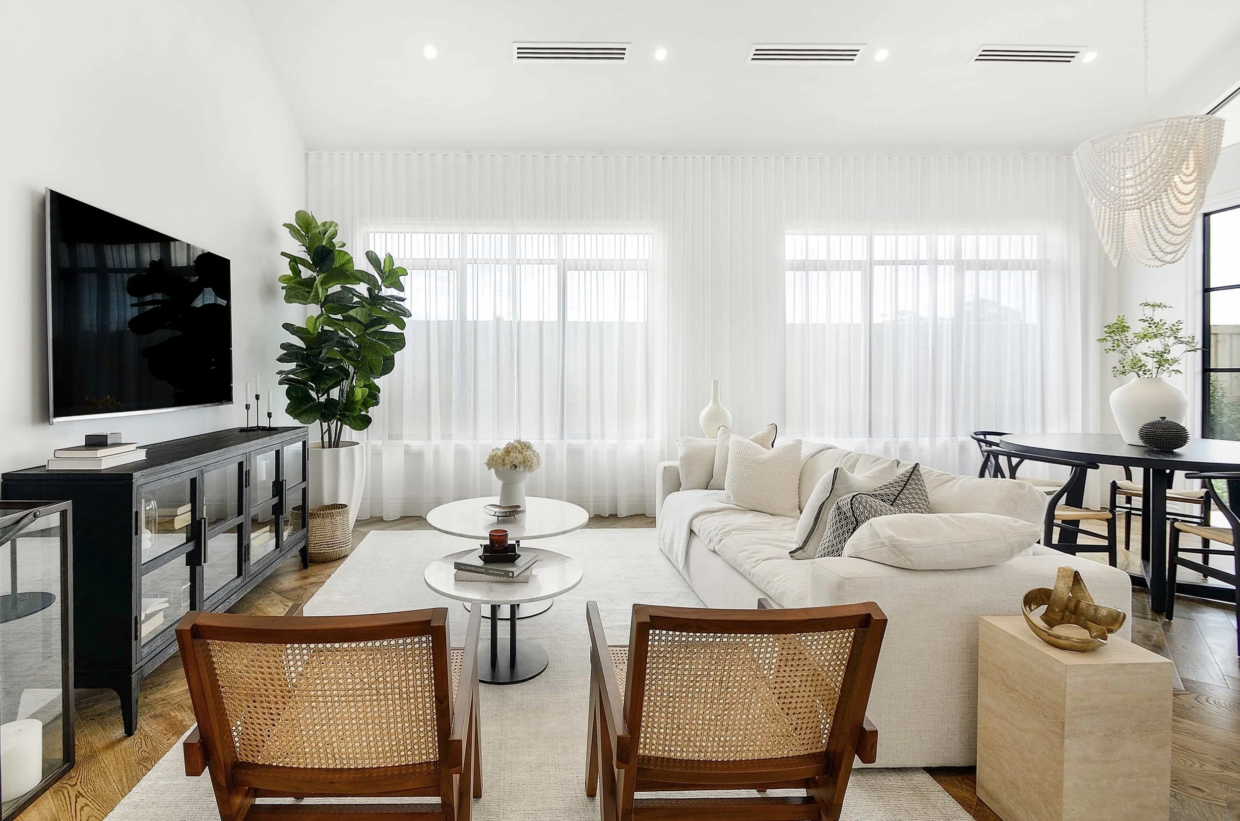 Bright, modern living room with white walls, large windows with sheer curtains, and wooden flooring. Features include a white sofa with decorative pillows, a round coffee table, a large potted plant, and a black console table with candles and books.