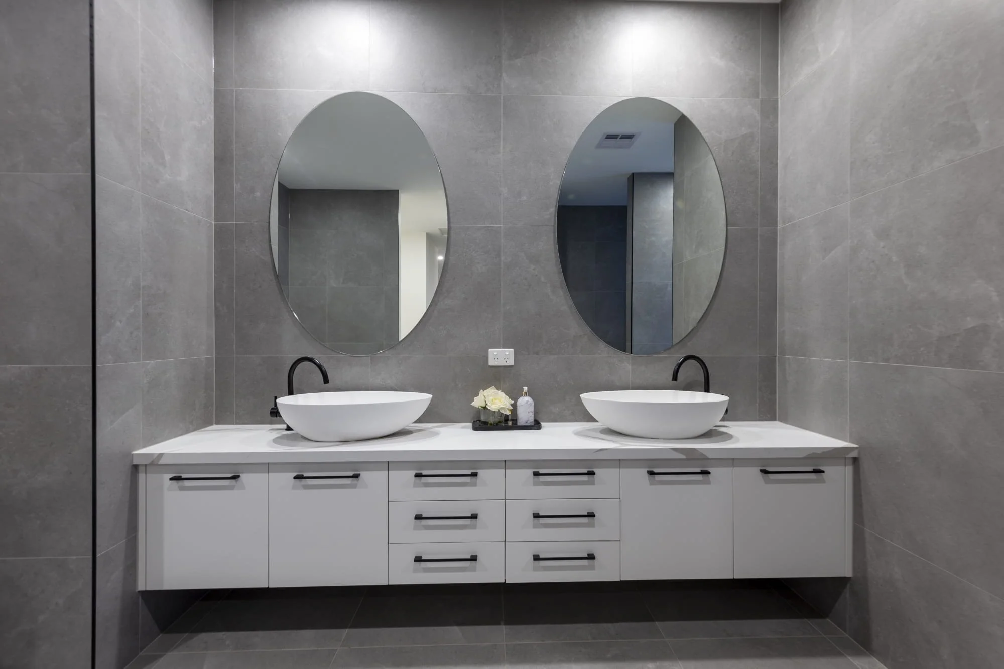 A modern floating ensuite vanity with a white benchtop, two basins, black faucets, and two large oval mirrors. There are small decorative items, including a flower bouquet, on the counter.