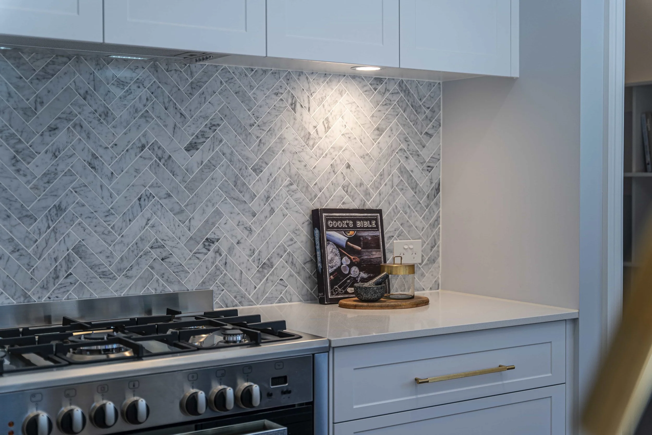 A section of a modern kitchen with white cabinets featuring gold handles and a white stone benchtop. A stainless steel stove is partially visible. The wall has a herringbone tile pattern.