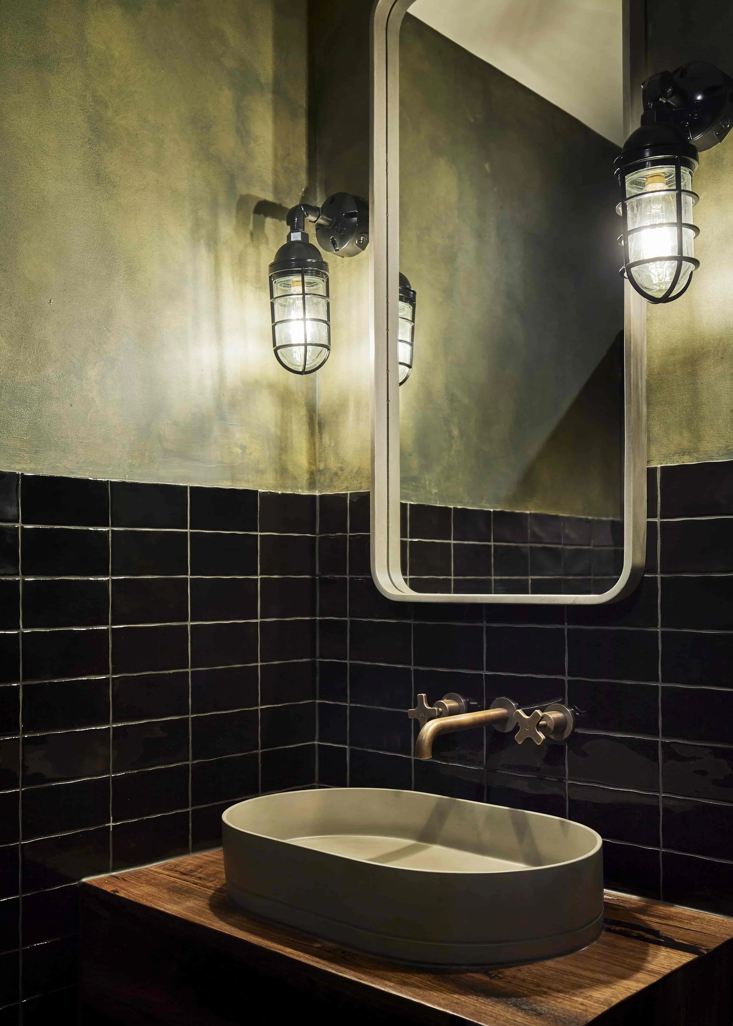 Moody bathroom with black tiled walls, a beige oval basin on a wooden vanity benchtop, and a framed mirror with vintage style wall lights on either side.