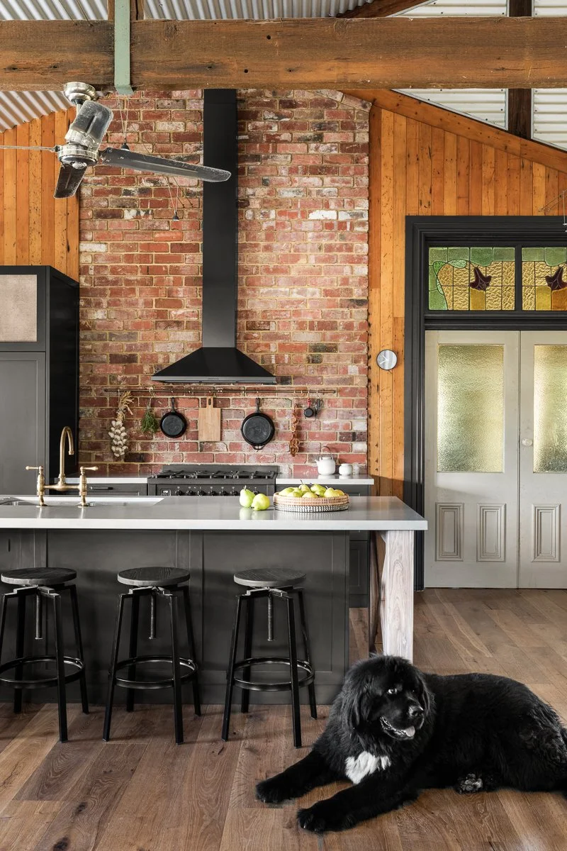 A rustic kitchen in a barn style home with exposed brick feature cladding, dark joinery, and copper accents.